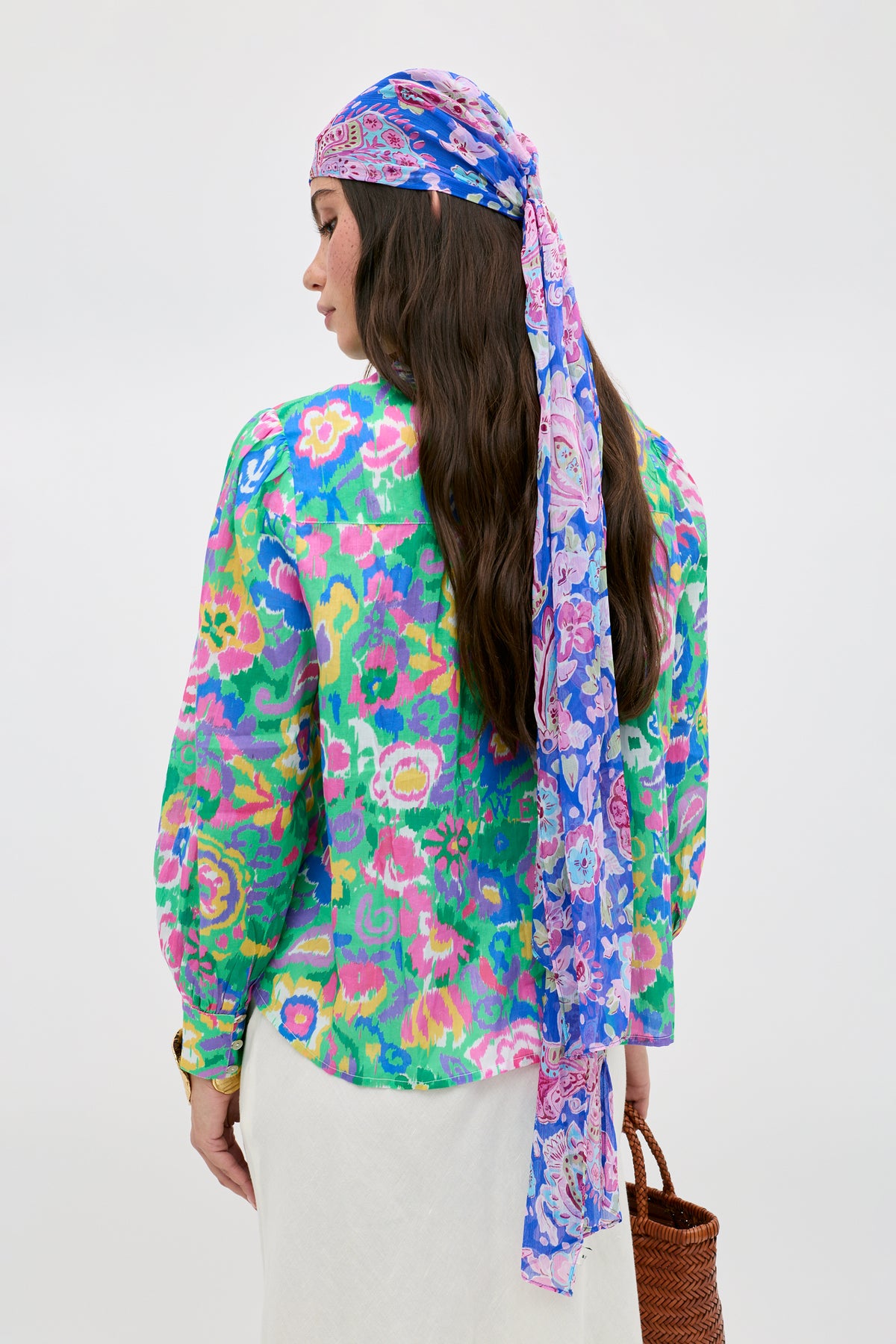 A woman facing away is wearing the Blake - Ikat Bloom Green blouse, a long purple and blue patterned headscarf, and a white skirt. She holds a woven bag in her left hand against a plain, light-colored background.