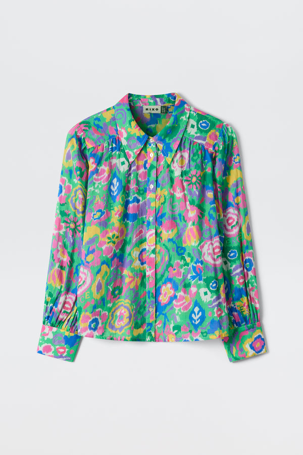 The "Blake - Ikat Bloom Green" is a long-sleeved blouse with a collar, showcasing a vibrant floral and abstract pattern in green, pink, blue, yellow, and purple on a light background.
