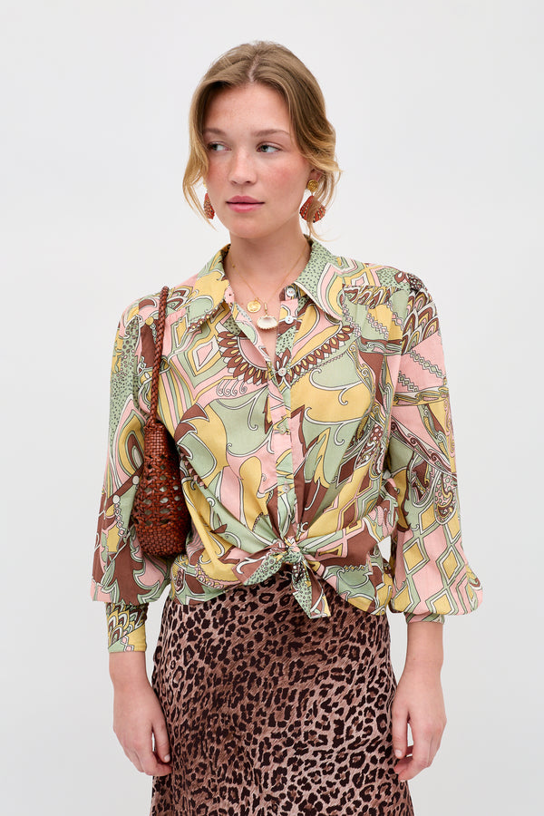 A woman wears the Blake - Bianca Abstract Green blouse, tied at the waist, with a brown animal print skirt and a brown woven shoulder bag. She stands against a plain light background, looking slightly to the side.