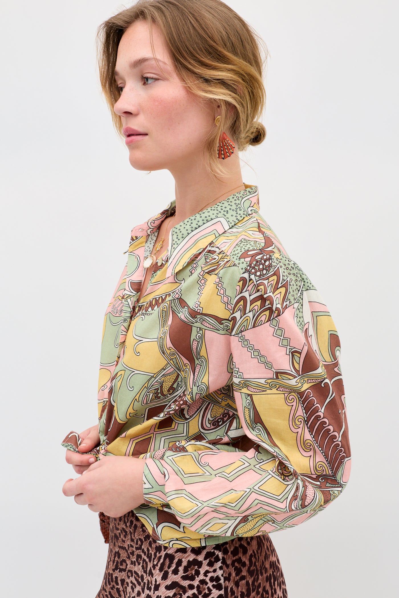 A woman stands in profile wearing the Blake - Bianca Abstract Green blouse with colorful geometric and abstract designs, paired with a leopard print skirt, red earrings, and her hair in a low, loose bun.