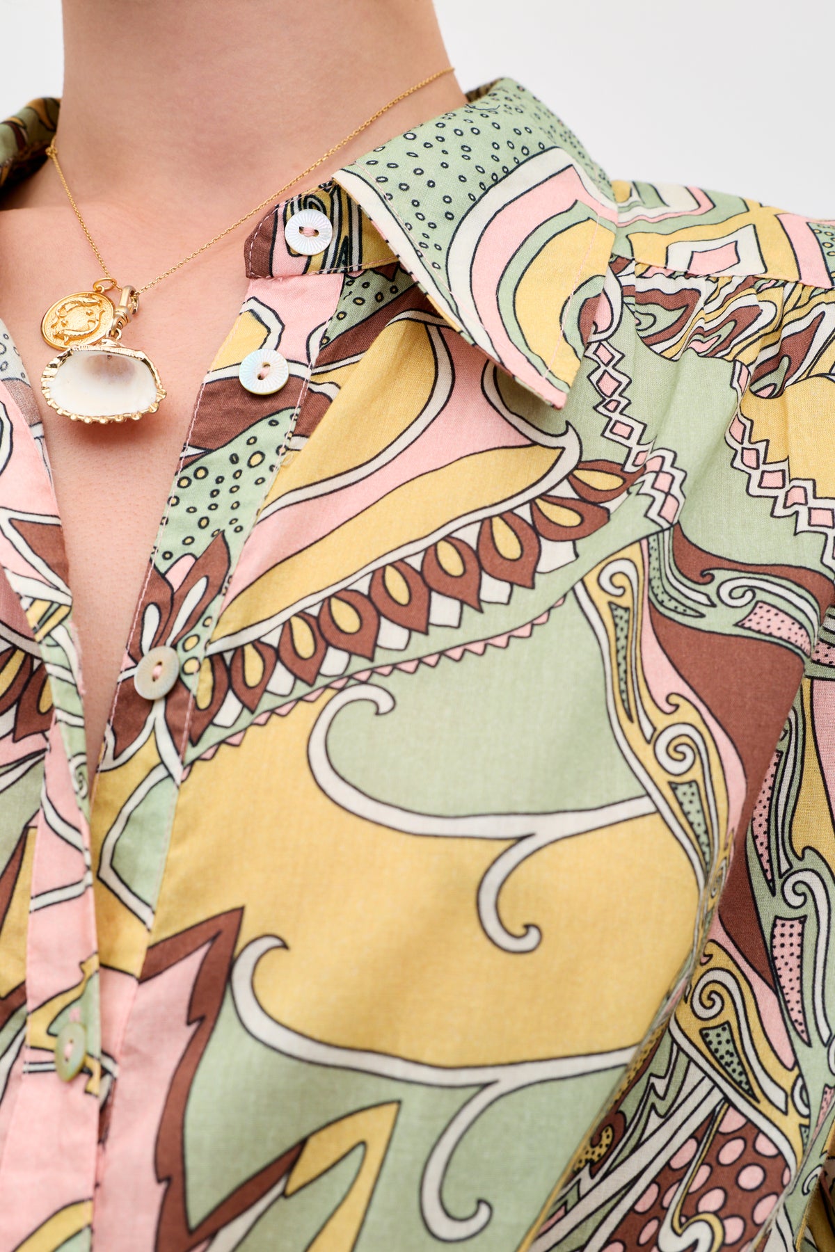 A close-up of a person wearing the Blake - Bianca Abstract Green shirt, featuring colorful swirling patterns in yellow, green, pink, and brown, paired with a gold necklace with a white shell pendant and round charm.