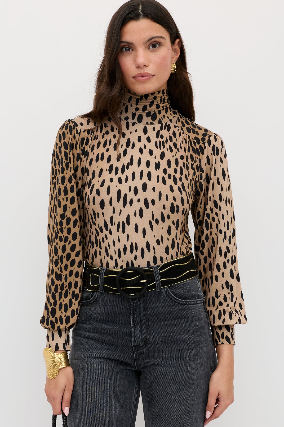A woman with long dark hair wears the Suki - Antelope Spot Black top, black jeans, a black belt with a gold buckle, and gold accessories, standing against a plain white background.