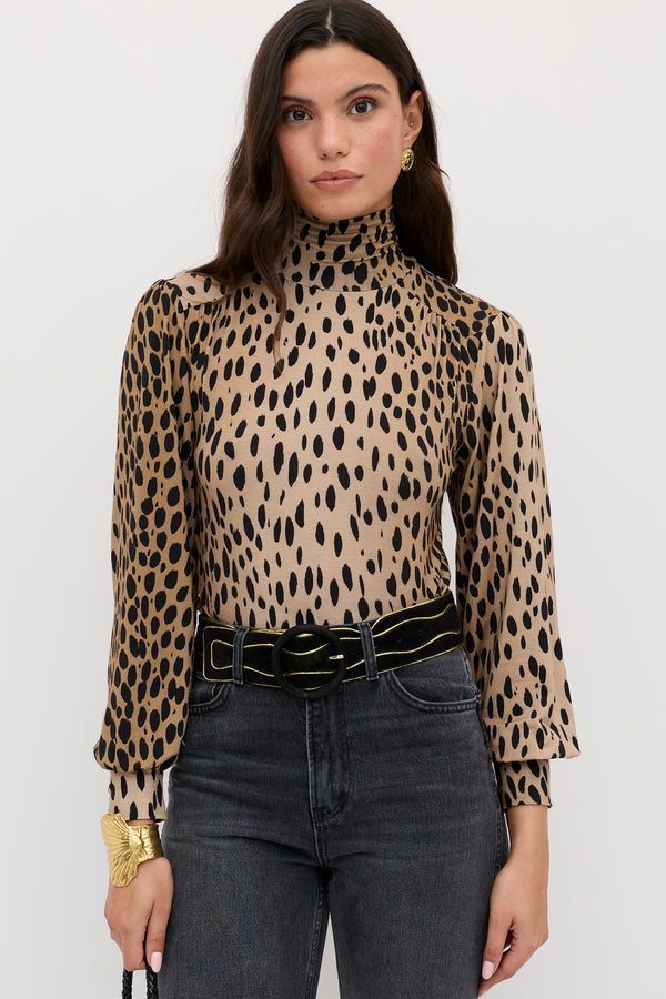 A woman with long dark hair wears the Suki - Antelope Spot Black top, black jeans, a black belt with a gold buckle, and gold accessories, standing against a plain white background.