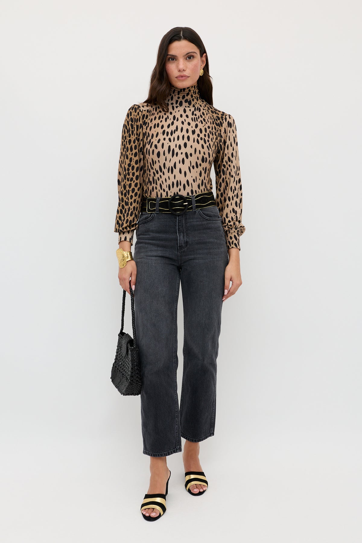 A woman stands against a white background wearing the Suki - Antelope Spot Black top with high-waisted black jeans, a black belt, black heeled sandals, and holding a black textured handbag.
