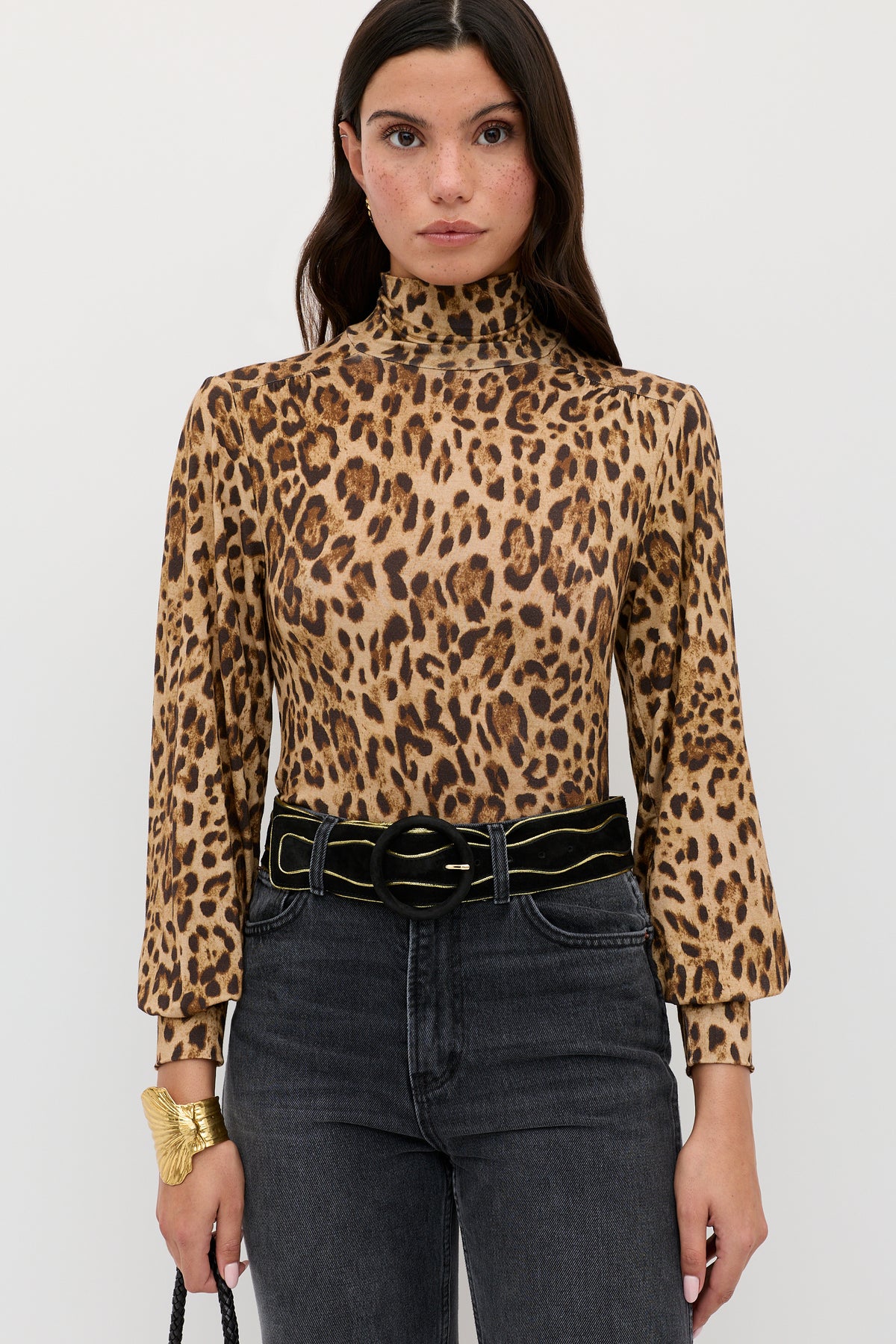Woman wearing the Suki - Mila Leopard Brown turtleneck layering top, black high-waisted jeans, and a black belt with a gold buckle. She has long, dark hair and holds a small black bag.