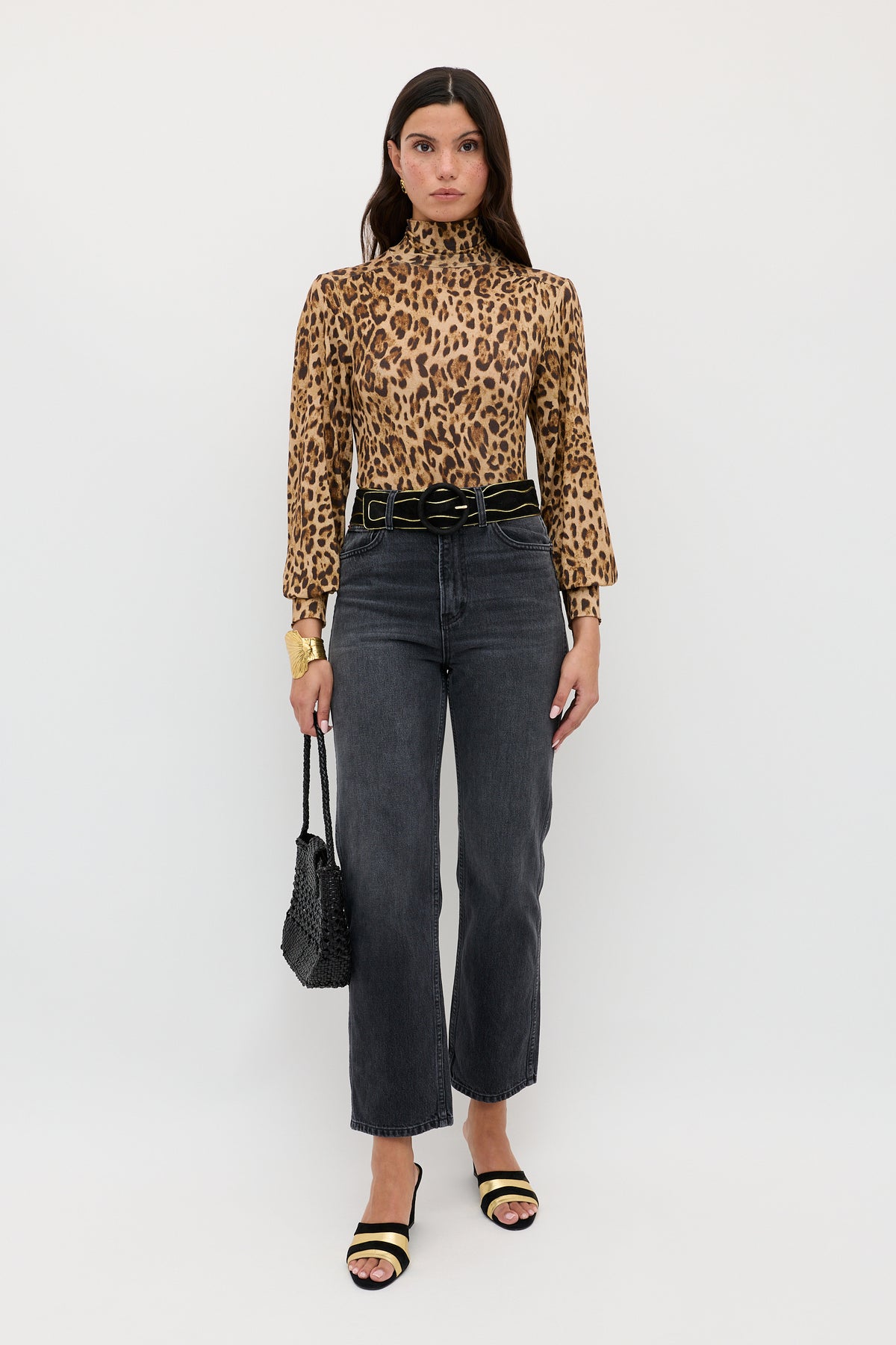 A woman stands against a white background, wearing the Suki - Mila Leopard Brown turtleneck layering top by RIXO, paired with high-waisted black jeans, a black belt, black sandals, and holding a black textured purse.