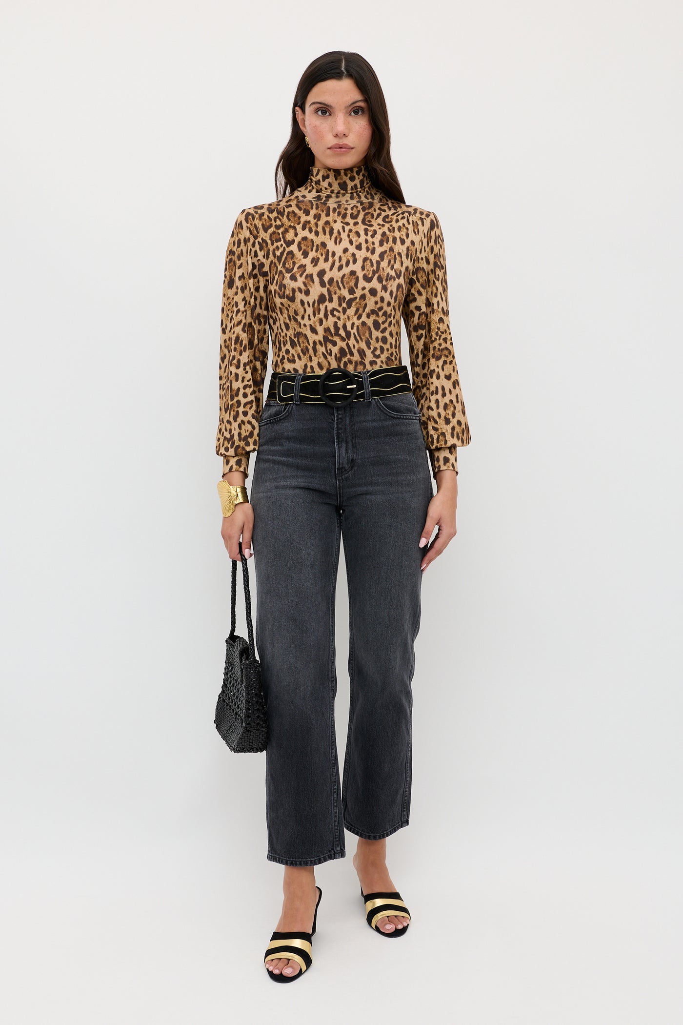 A woman stands against a white background, wearing the Suki - Mila Leopard Brown turtleneck layering top by RIXO, paired with high-waisted black jeans, a black belt, black sandals, and holding a black textured purse.