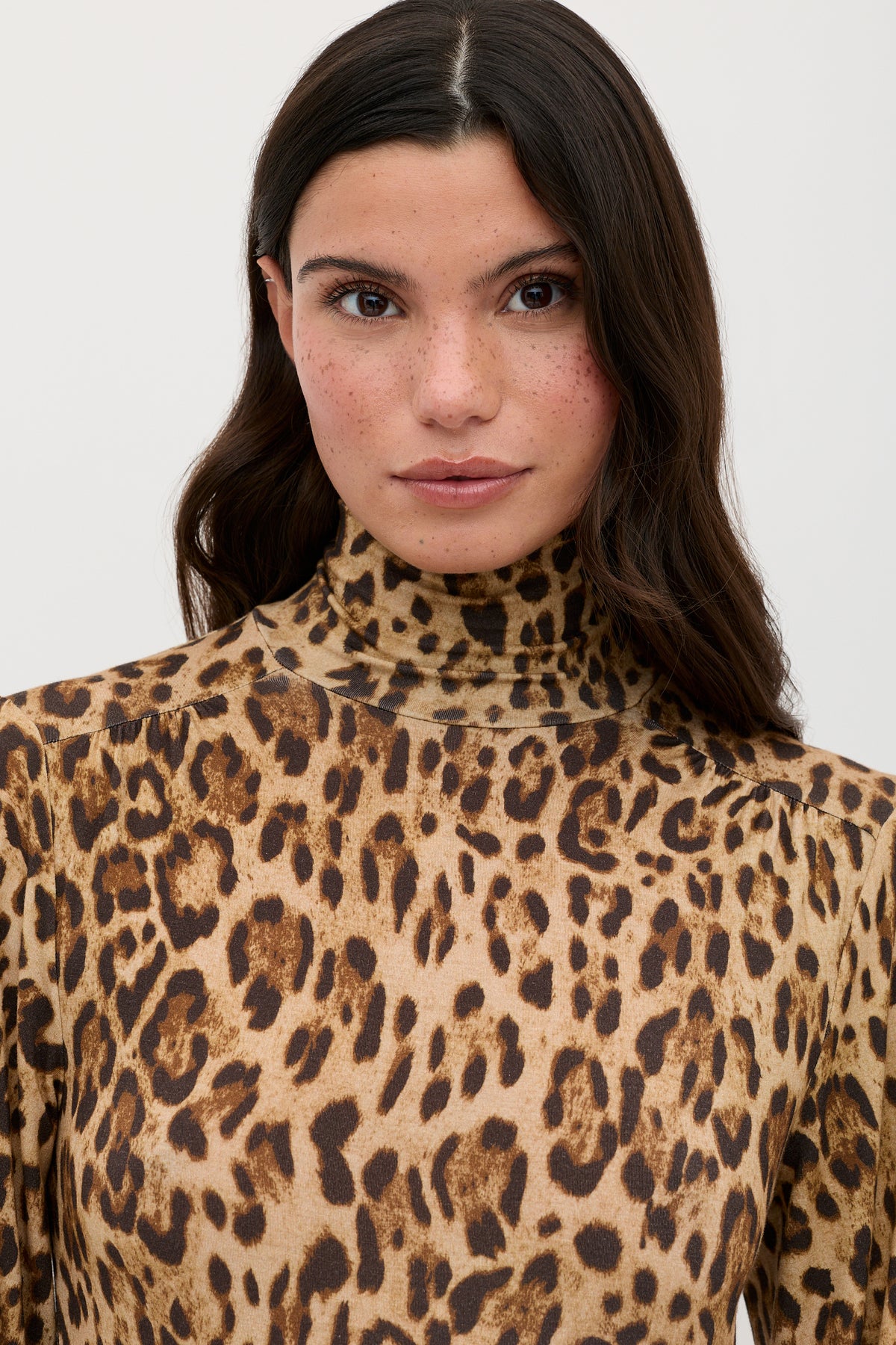 A woman with long dark hair, wearing the Suki - Mila Leopard Brown high-neck, long-sleeve top from RIXO, featuring a brown and black leopard print pattern, stands against a plain light background.