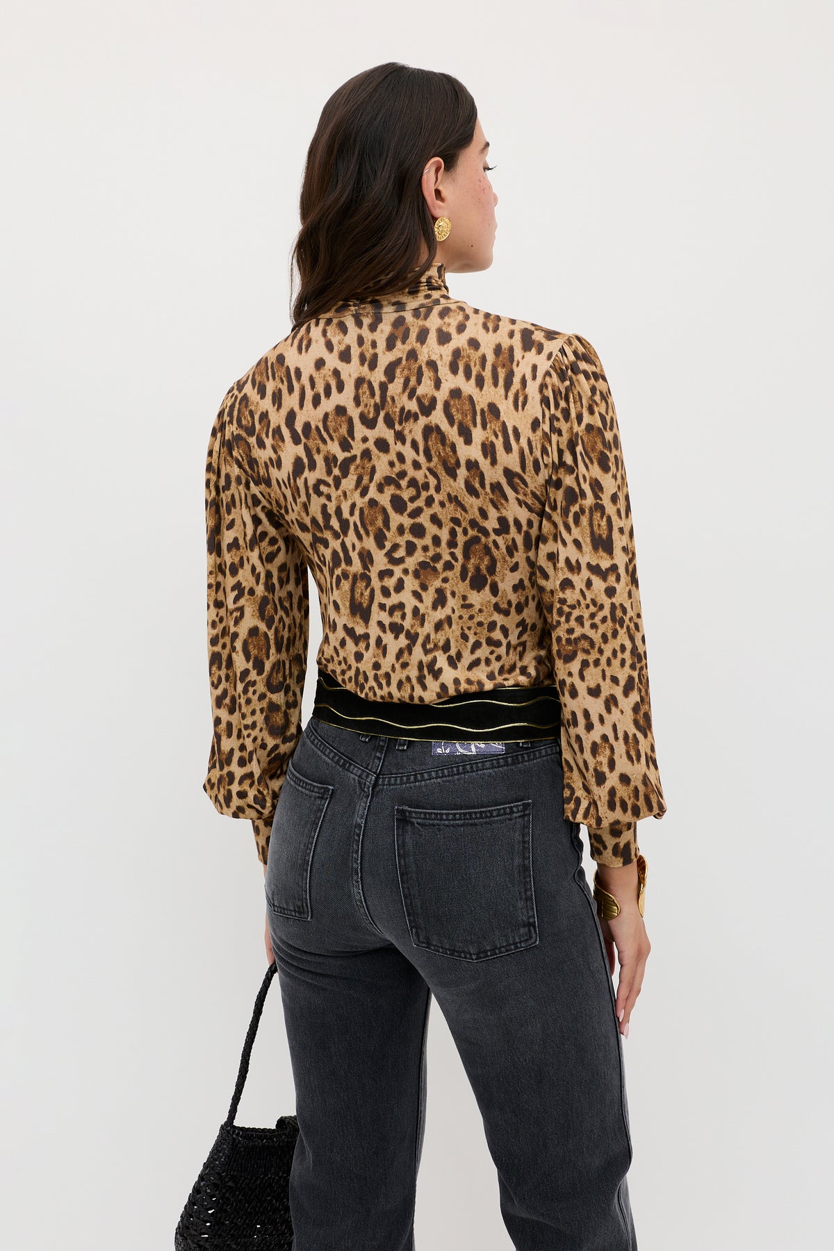 A woman with long dark hair, accessorized with gold hoop earrings, is shown from the back wearing the Suki - Mila Leopard Brown blouse by RIXO. She pairs it with high-waisted black jeans, a black belt, and holds a textured black bag against a plain white background.