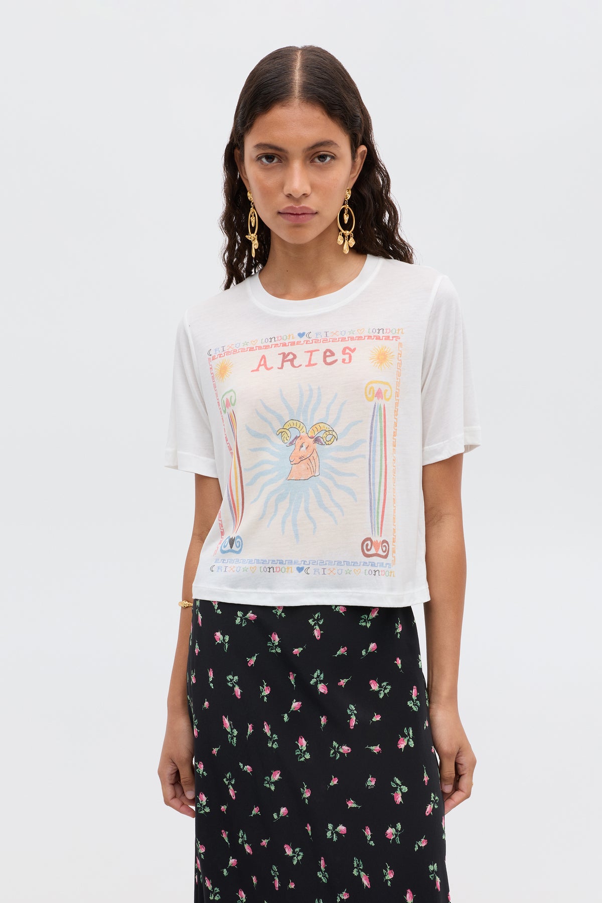 A woman with long, wavy dark hair wears gold hoop earrings and the Ria - Aries white T-shirt with a colorful Aries design and cropped hem, paired with a black skirt with a pink floral print, standing against a plain light background.