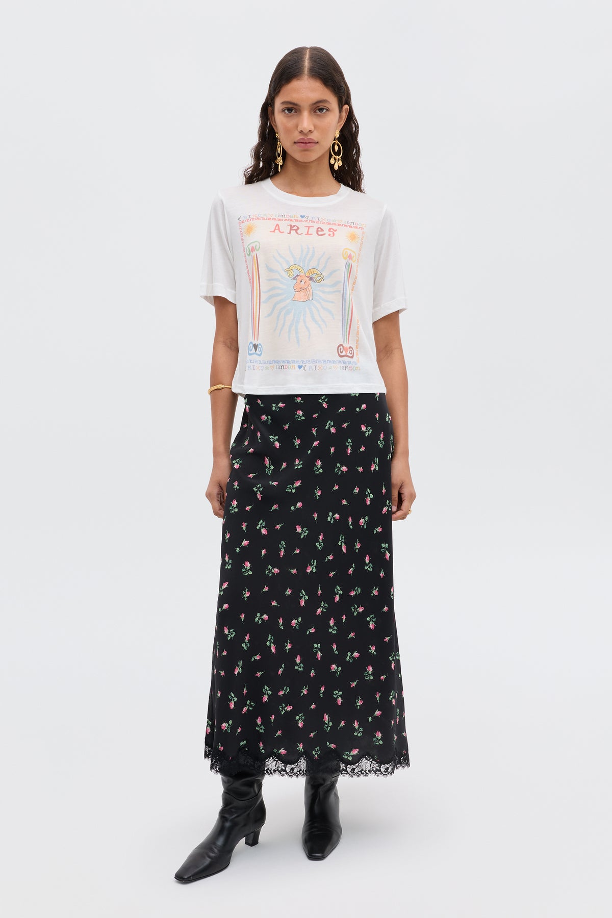 A woman with long, wavy hair and a neutral expression faces forward, wearing the white Ria - Aries T-shirt, a long black floral lace-trim skirt, black boots, and gold hoop earrings.