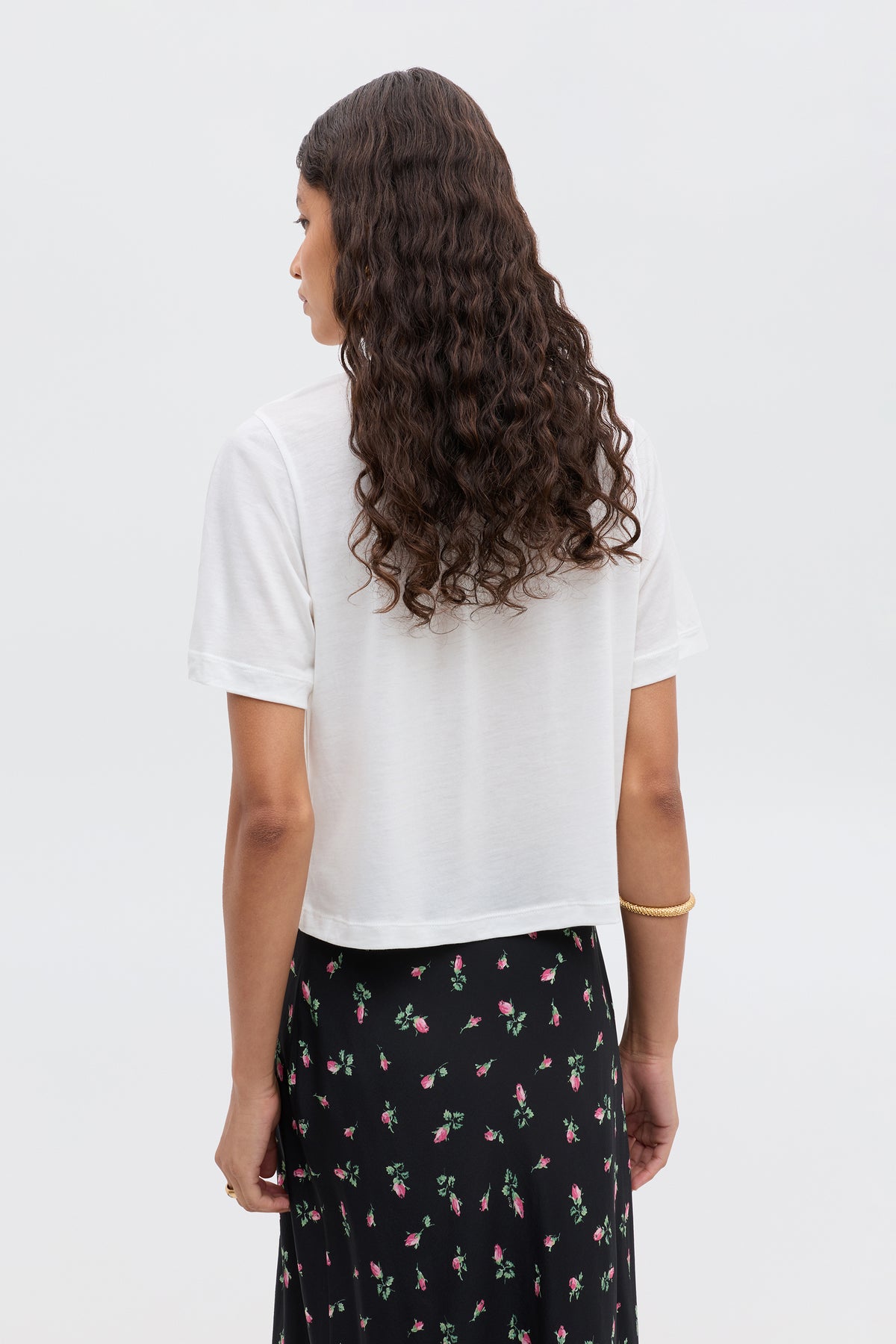 A woman with long, curly hair stands with her back to the camera, wearing the Ria - Aries cropped white T-shirt and a black skirt with a subtle floral pattern.