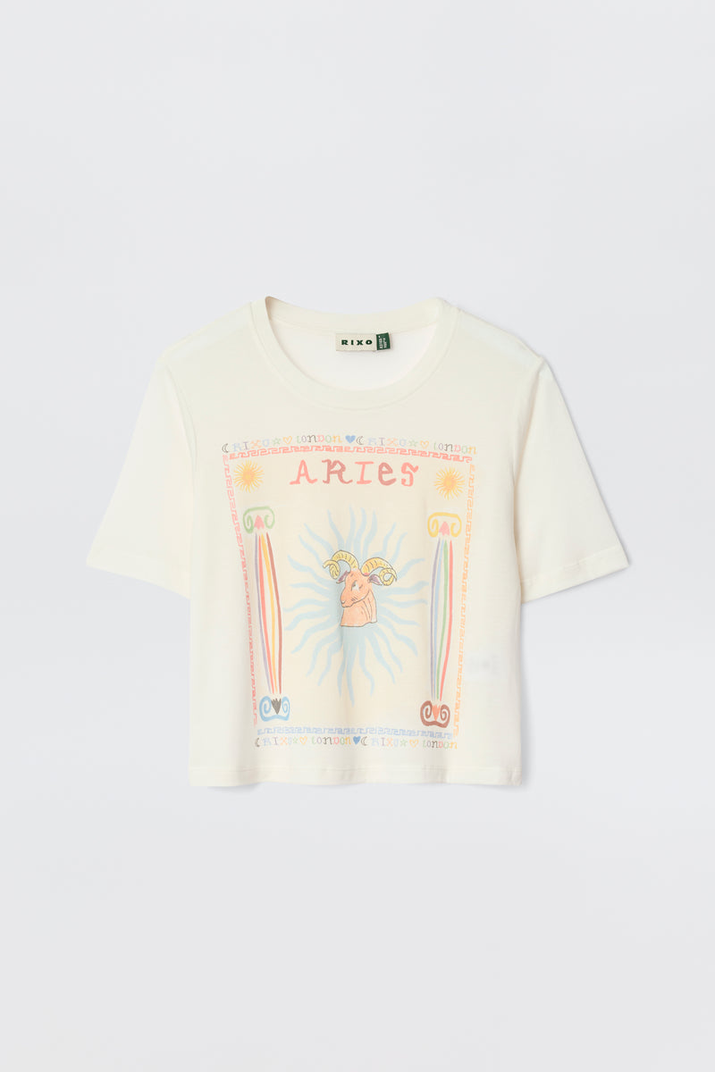 The Ria - Aries white short-sleeve T-shirt displays a vibrant Aries graphic with "ARIES" at the top, a central ram, sun rays, and pink, orange, and blue astrological symbols.