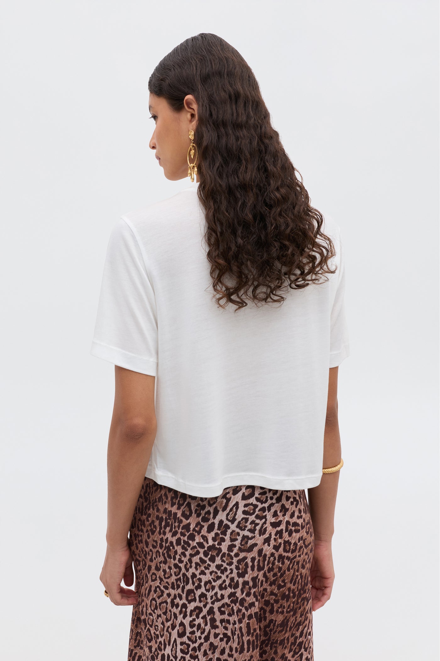 A woman with long, wavy dark hair is seen from behind in a white cropped tee and the brown leopard-print Ria - Taurus skirt by RIXO, accessorized with gold earrings and bracelets against a plain light background.