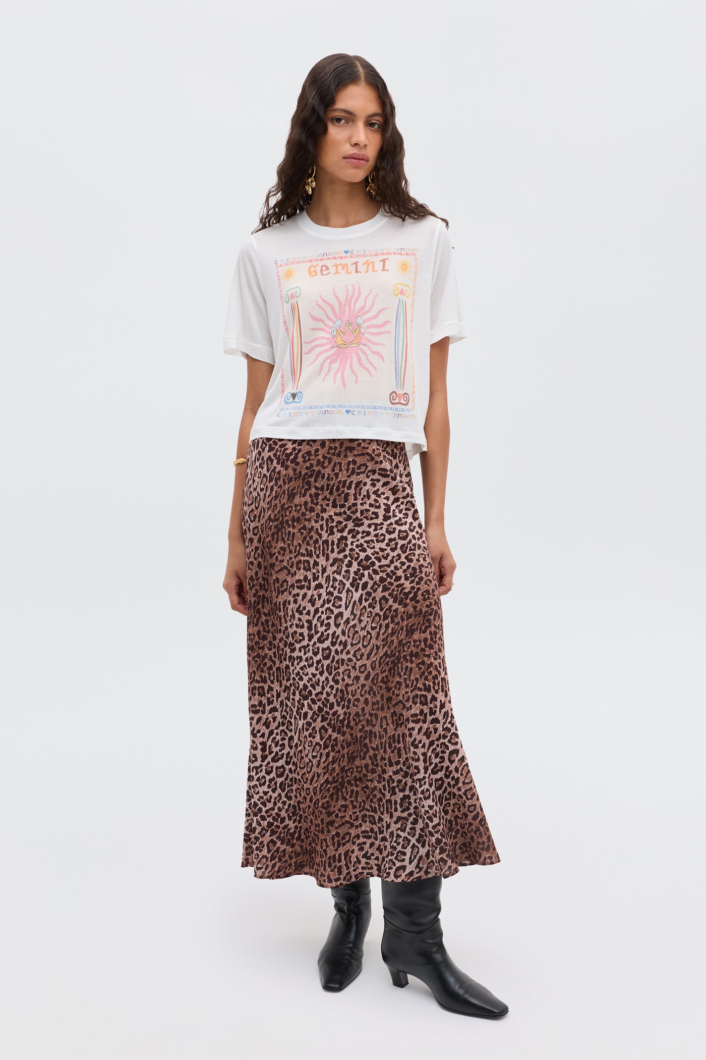 A woman stands against a plain background wearing the Ria - Gemini T-shirt in white, styled with a brown leopard print midi skirt and black pointed-toe boots.