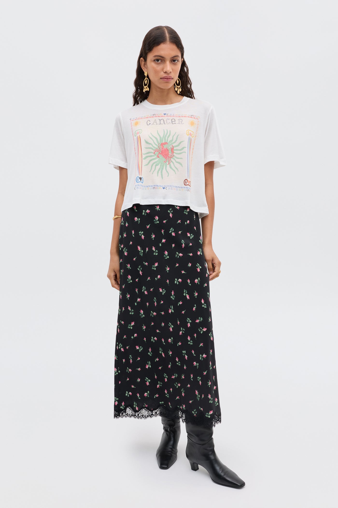 A woman poses against a plain background wearing the white Ria - Cancer T-shirt, a long black floral RIXO skirt with a lace hem, gold earrings, and black boots.