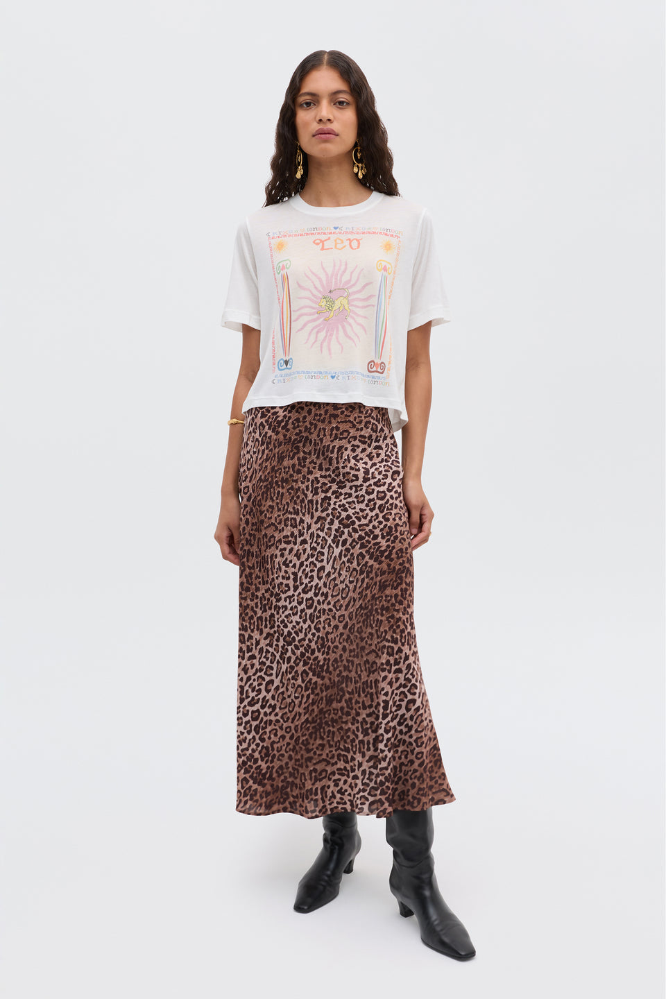 A woman with long wavy hair and a neutral expression stands against a plain background, wearing a white graphic tee, the Ria - Leo skirt with cropped hem, black knee-high boots, and gold earrings and bracelet.