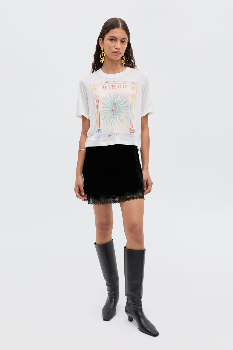A woman stands against a plain white background wearing the Ria - Virgo graphic t-shirt, a black lace-trim mini skirt, black knee-high boots, gold hoop earrings, and a gold bracelet.