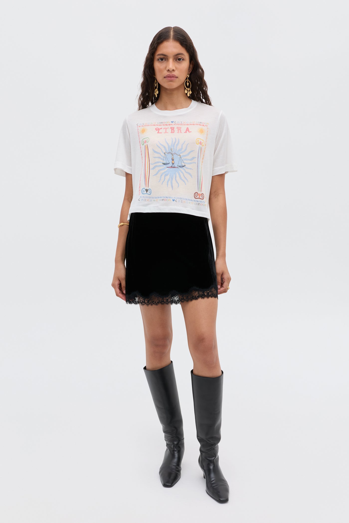 A woman with long, wavy hair and gold earrings stands against a white background, wearing the Ria - Libra white T-shirt with a colorful illustration, a lace-trimmed RIXO skirt with cropped hem, and black knee-high boots.
