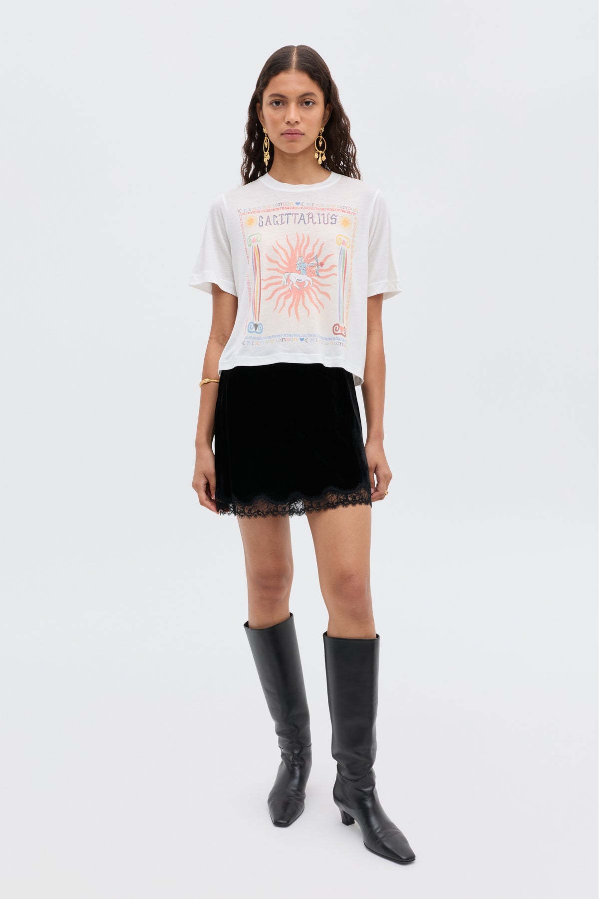 A woman with long wavy hair and gold earrings stands against a plain background wearing the Ria - Sagittarius white t-shirt, a black RIXO skirt with lace trim and cropped hem, and black knee-high boots.