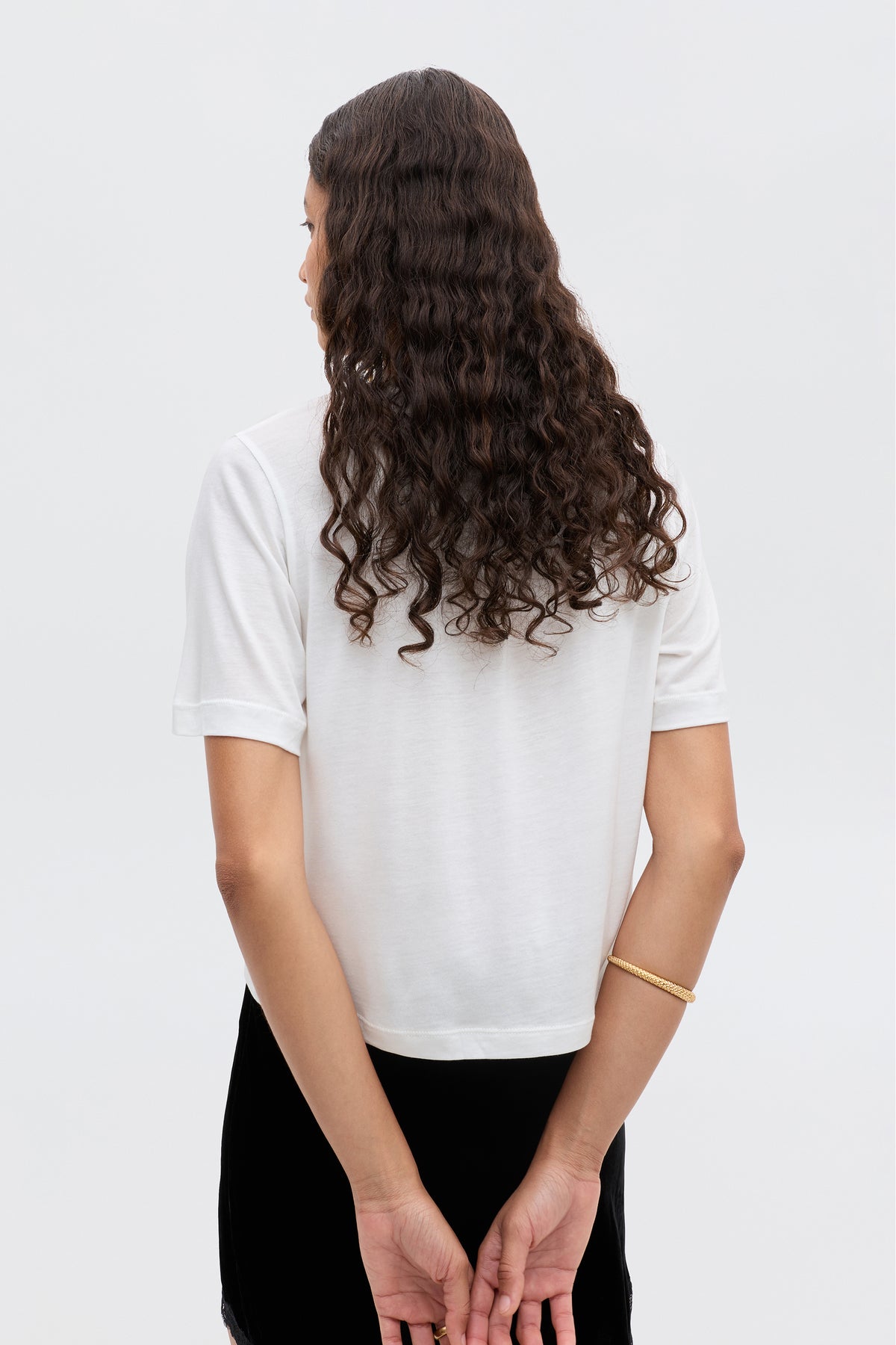 A person with long, curly hair models the Ria - Sagittarius white short-sleeve cropped shirt, standing with their back to the camera, hands behind their back, wearing a gold bracelet against a plain light background.