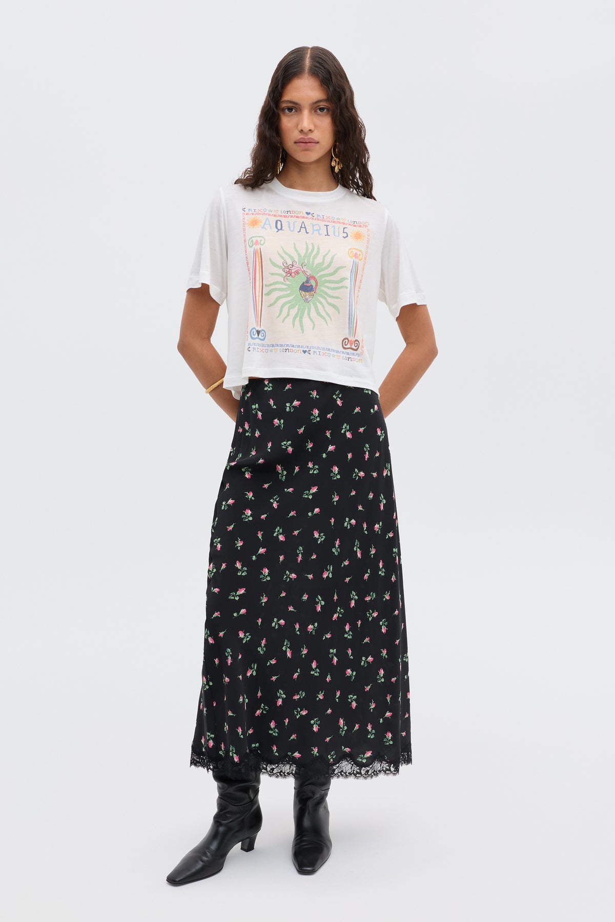 A woman poses against a white background wearing the Ria - Aquarius white illustration t-shirt with a cropped hem, a long black skirt with floral lace trim, and black boots.