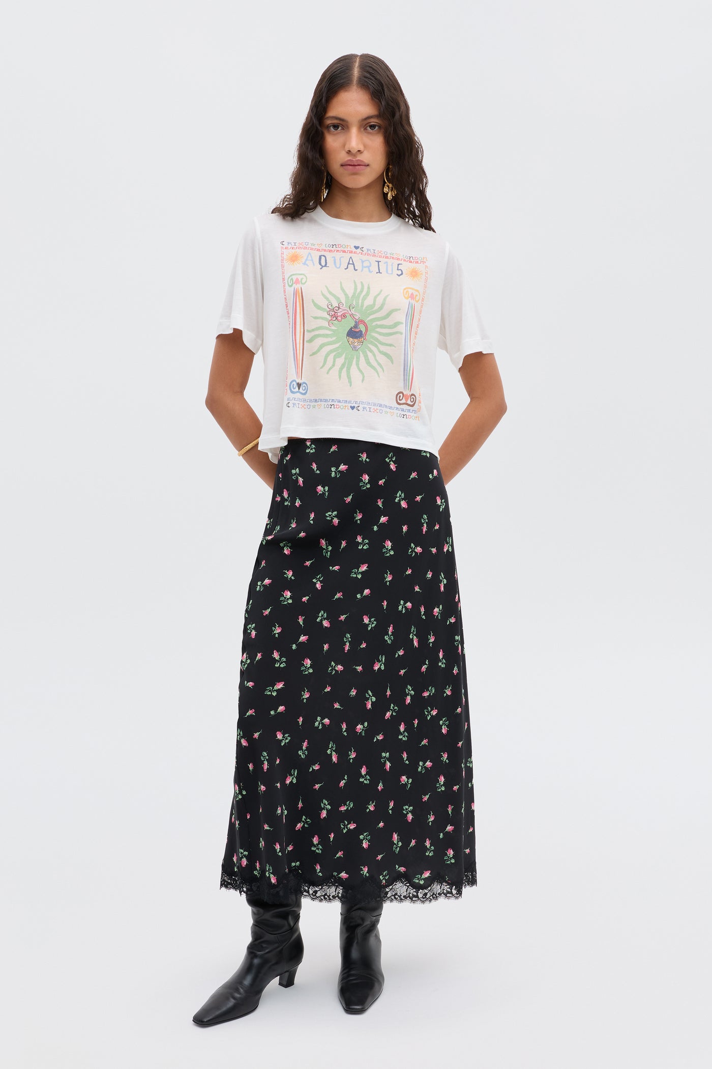 A woman poses against a white background wearing the Ria - Aquarius white illustration t-shirt with a cropped hem, a long black skirt with floral lace trim, and black boots.