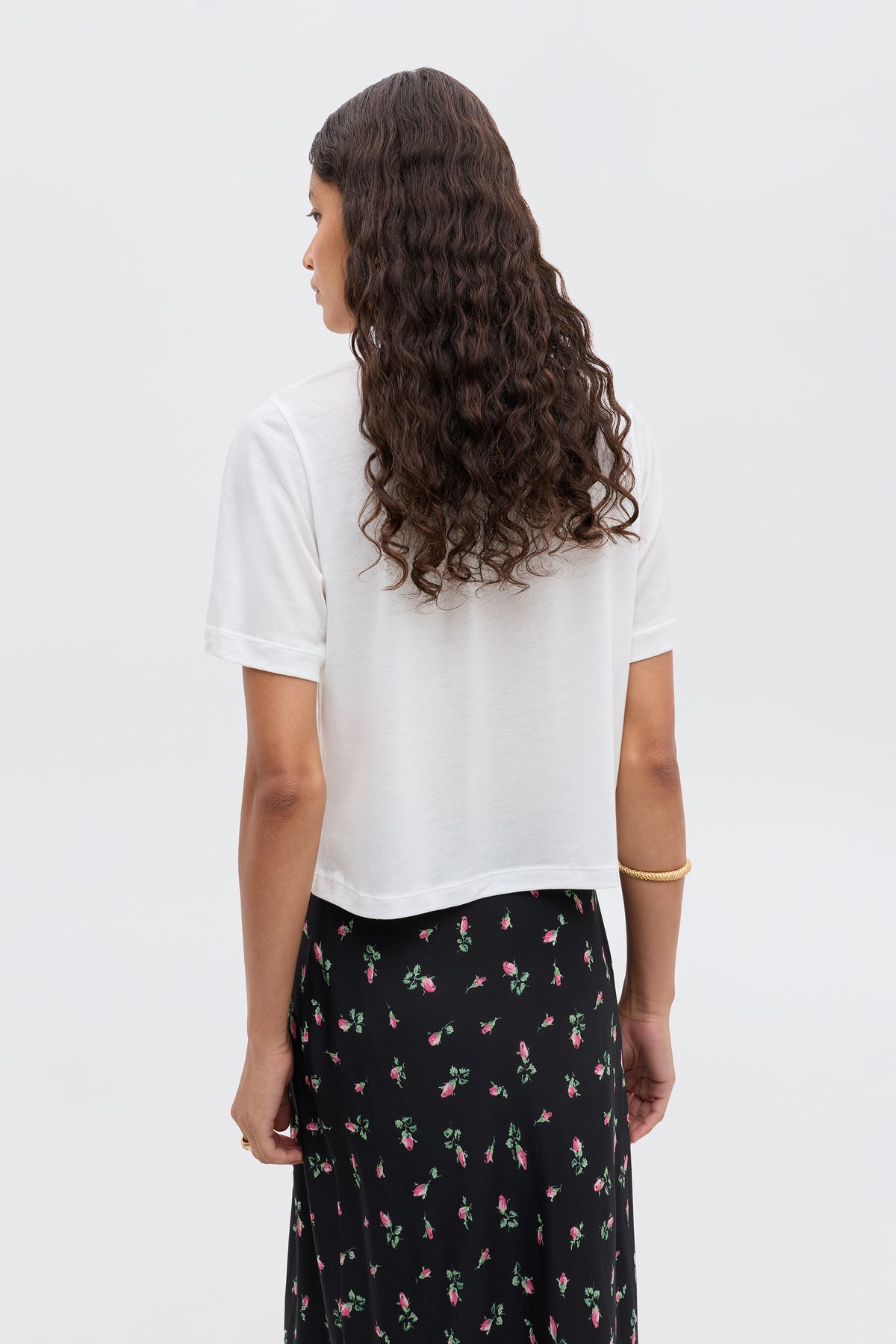 A person with long curly hair, in a white short-sleeve shirt and the black RIXO "Ria - Aquarius" skirt with a cropped hem, is standing with their back to the camera against a plain light background.