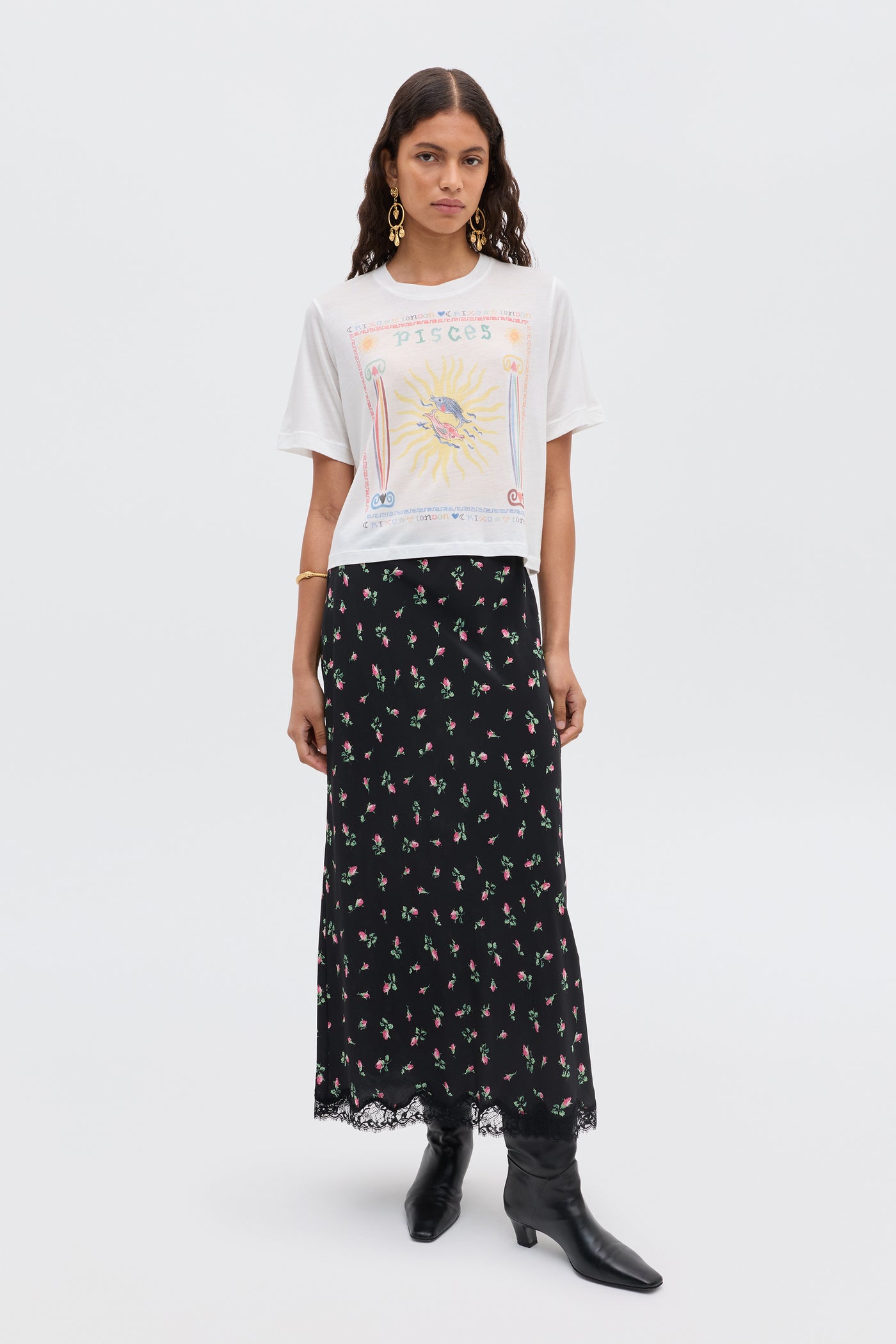 A woman stands against a plain background wearing the Ria - Pisces white cropped hem T-shirt with a Pisces illustration, a long black skirt with lace trim, black boots, and gold earrings.
