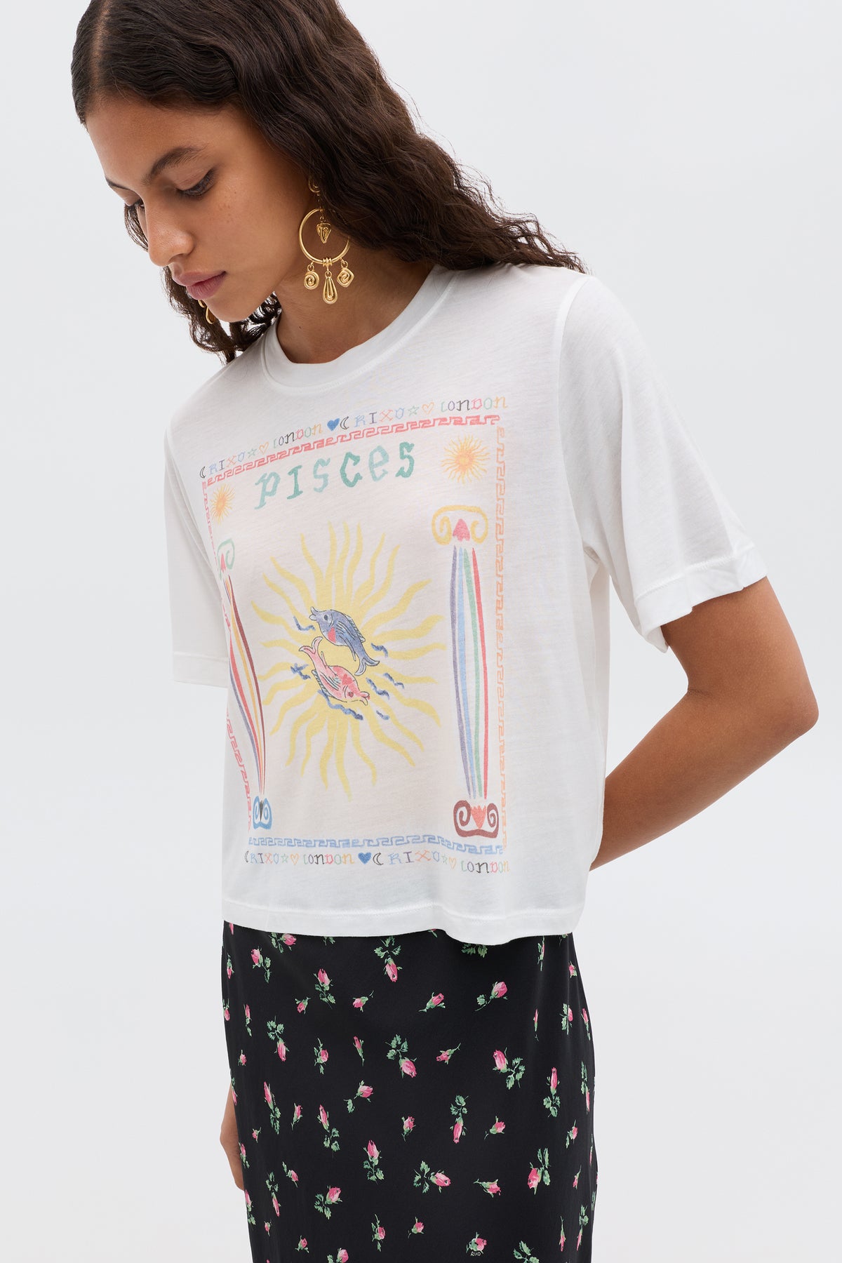 A woman with long wavy hair and large gold earrings wears the Ria - Pisces white cropped hem T-shirt featuring a colorful Pisces illustration, paired with a black floral skirt, against a plain light background.
