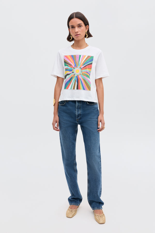 A woman stands against a plain background wearing the Ria- Casa Soleil Multi T-shirt, featuring a colorful radiating geometric design, paired with cropped blue jeans, woven beige flats, gold earrings, and a gold bracelet.