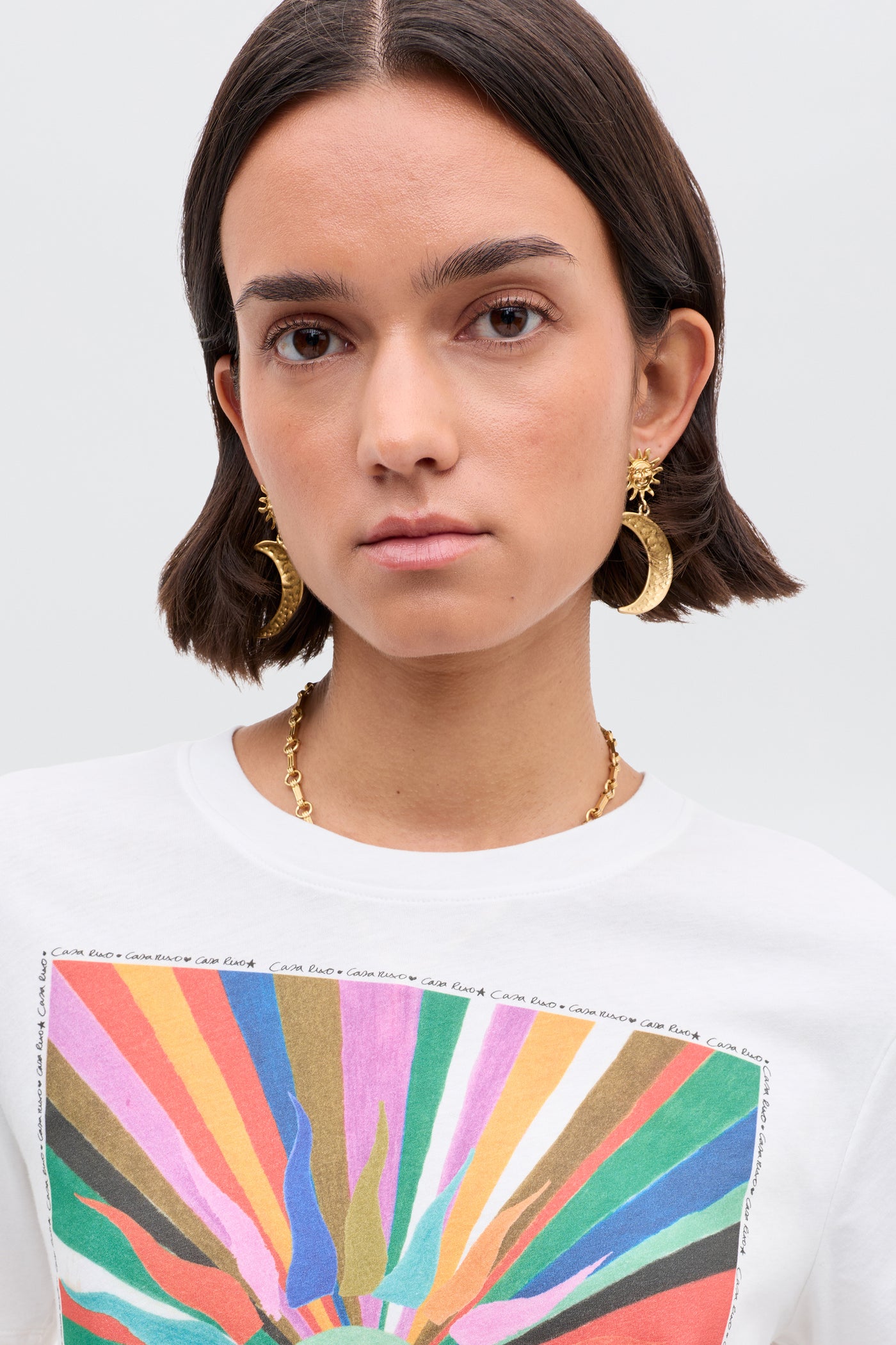 A woman with short dark hair, wearing gold moon-shaped earrings, a gold necklace, and a Casa RIXO Ria- Casa Soleil Multi T-shirt with a cropped hem and colorful abstract sunburst pattern, looks at the camera neutrally.
