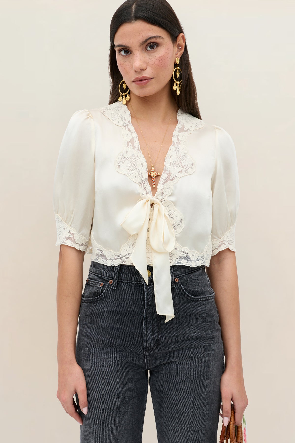 A woman wears the Amara - Champagne, a cream lace short-sleeve blouse with a front bow, paired with high-waisted dark jeans. She has long straight hair, gold earrings, and a neutral expression against a plain background.