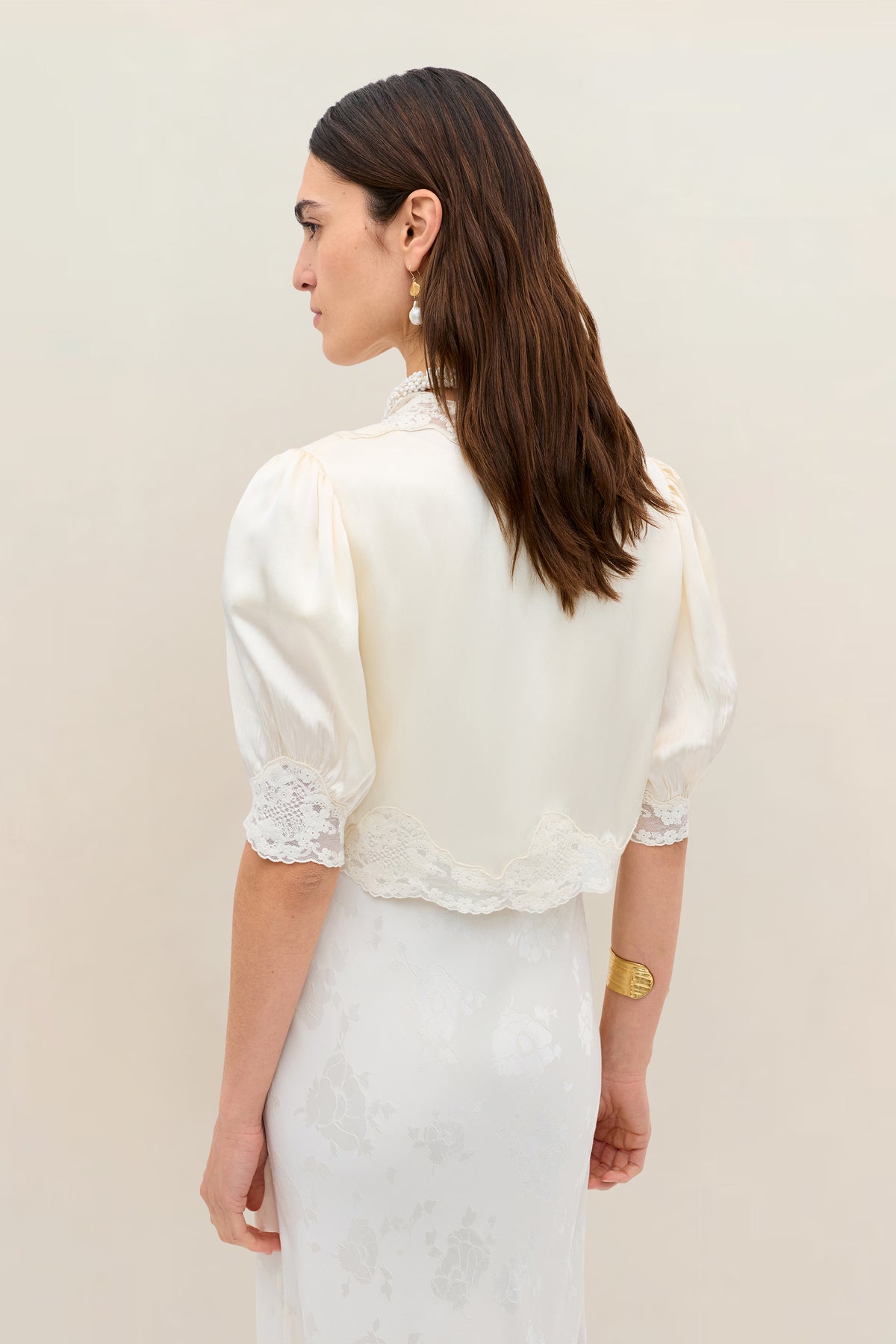 A woman with long brown hair stands with her back to the camera, wearing the Amara - Champagne top with lace trim over a silk-satin dress. She accessorizes with gold earrings and a bracelet, set against a neutral background.