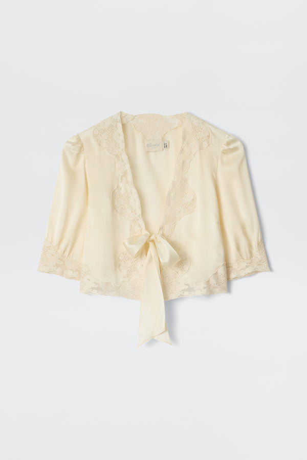 The Amara - Champagne is a light cream satin blouse with delicate lace trim, three-quarter sleeves, and a deep V-neckline that ties at the front—ideal for bridal looks or any elegant occasion.