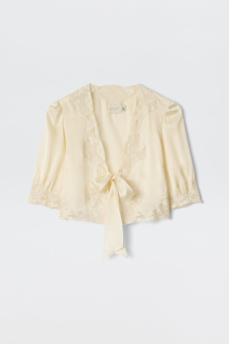 The Amara - Champagne is a light cream satin blouse with delicate lace trim, three-quarter sleeves, and a deep V-neckline that ties at the front—ideal for bridal looks or any elegant occasion.