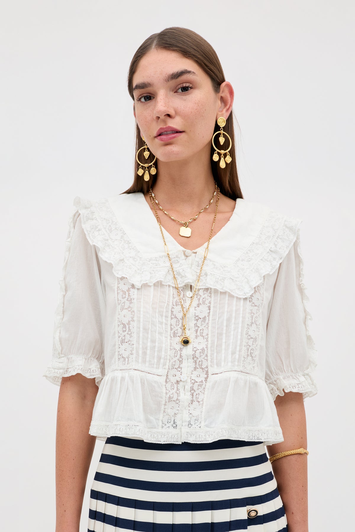 A woman wears the Ellery - White vintage-inspired blouse with lace details and a ruffled collar, paired with a navy and white striped skirt, gold drop earrings, layered necklaces, and a bracelet against a plain background.