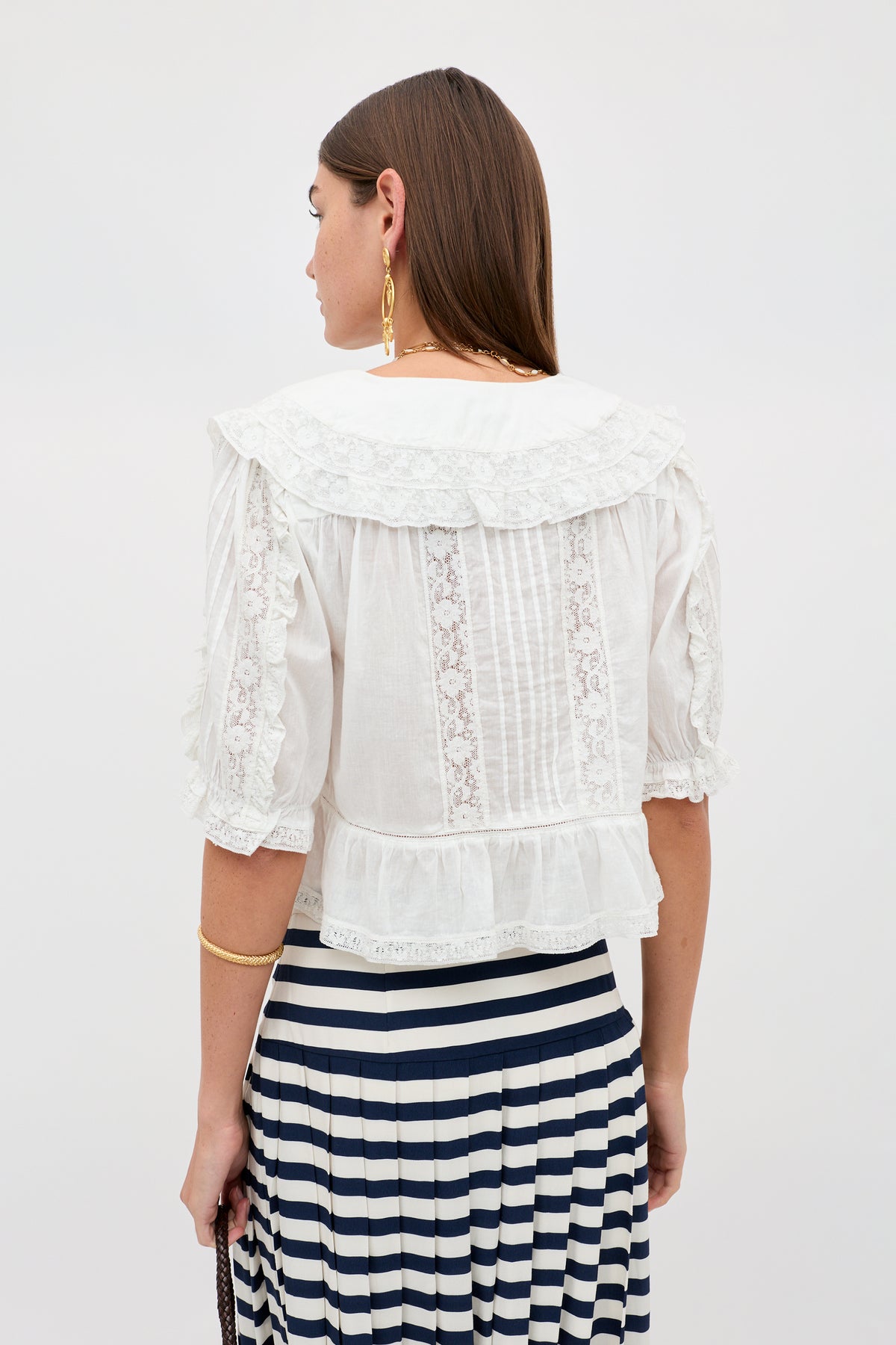 A woman with straight brown hair wears the Ellery - White blouse, a vintage-inspired white cotton voile top with ruffles and a sailor collar. Facing away, she shows off its back, gold jewelry, and a black-and-white striped skirt.