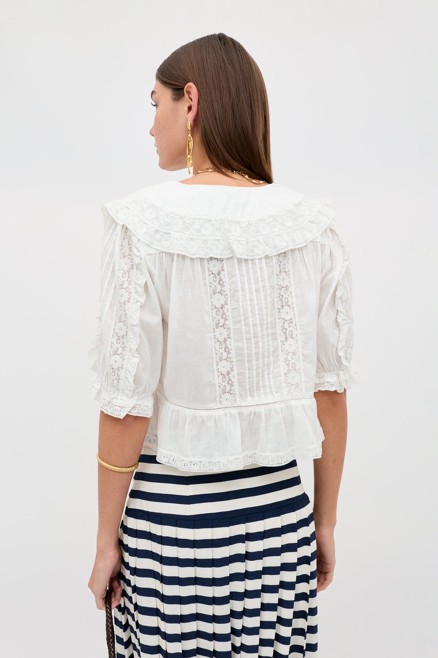 A woman with straight brown hair wears the Ellery - White blouse, a vintage-inspired white cotton voile top with ruffles and a sailor collar. Facing away, she shows off its back, gold jewelry, and a black-and-white striped skirt.