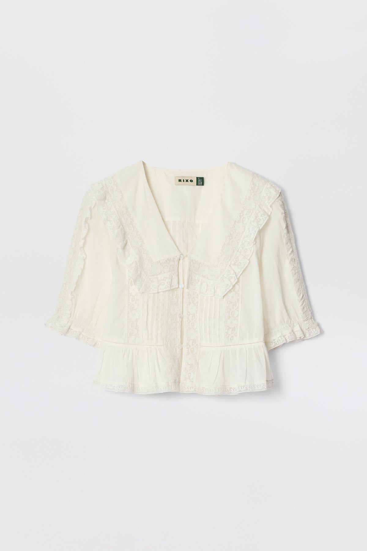 The Ellery - White is a cream-colored, vintage-inspired blouse made from lightweight cotton voile with a wide ruffled lace collar, short puff sleeves, front pleats, and delicate lace trim, shown on a plain white background.