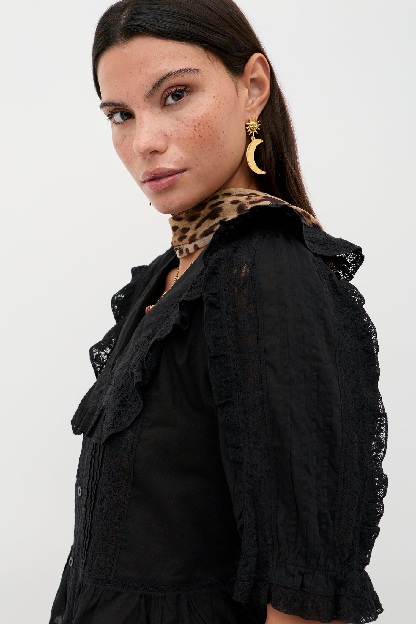 A woman with long dark hair and freckles wears the Ellery - Black vintage-inspired cotton button-down top, styled with a leopard print scarf and gold crescent moon earring, looking at the camera against a plain white background.