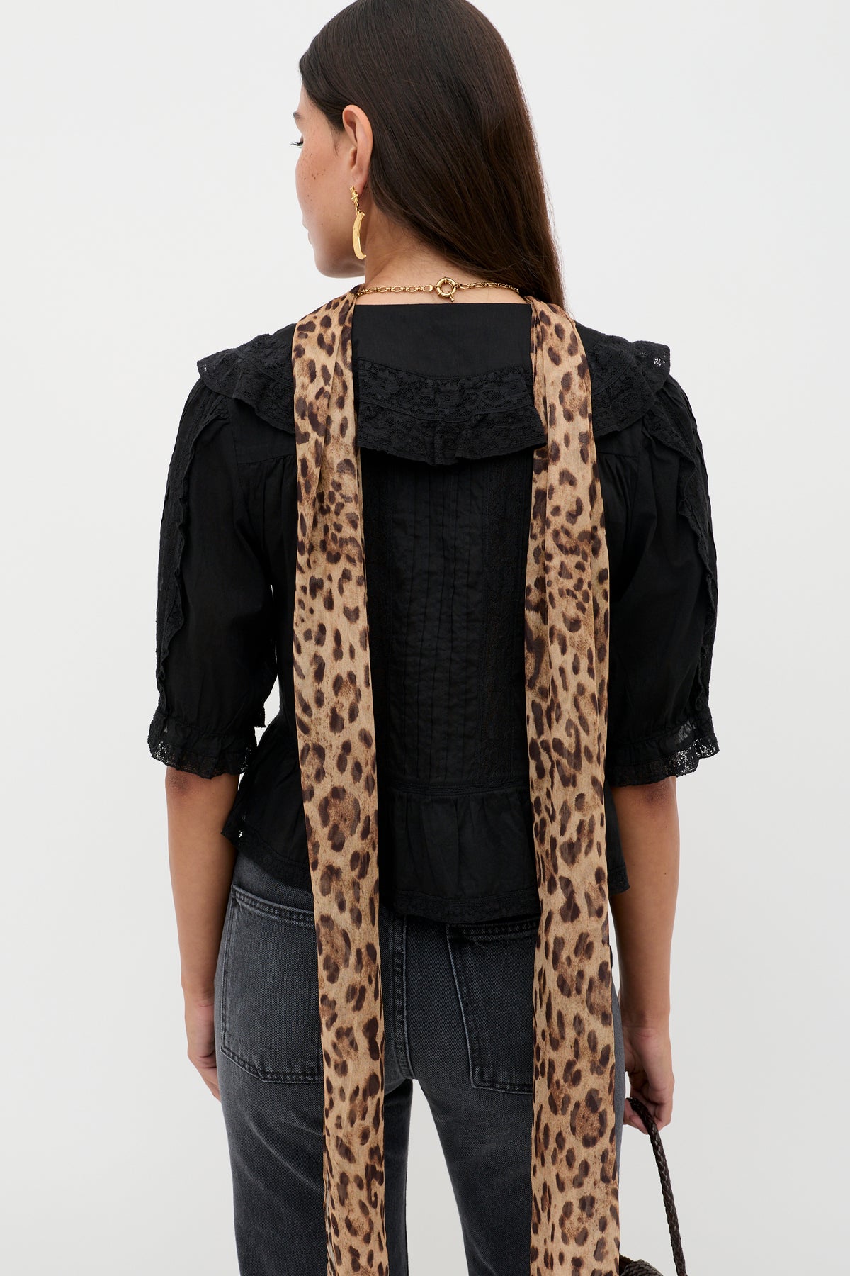 A woman with long dark hair stands facing away, wearing the Ellery - Black vintage-inspired cotton button-down top with ruffled collar and sleeves, paired with dark jeans, gold hoop earrings, and a draped leopard print scarf.
