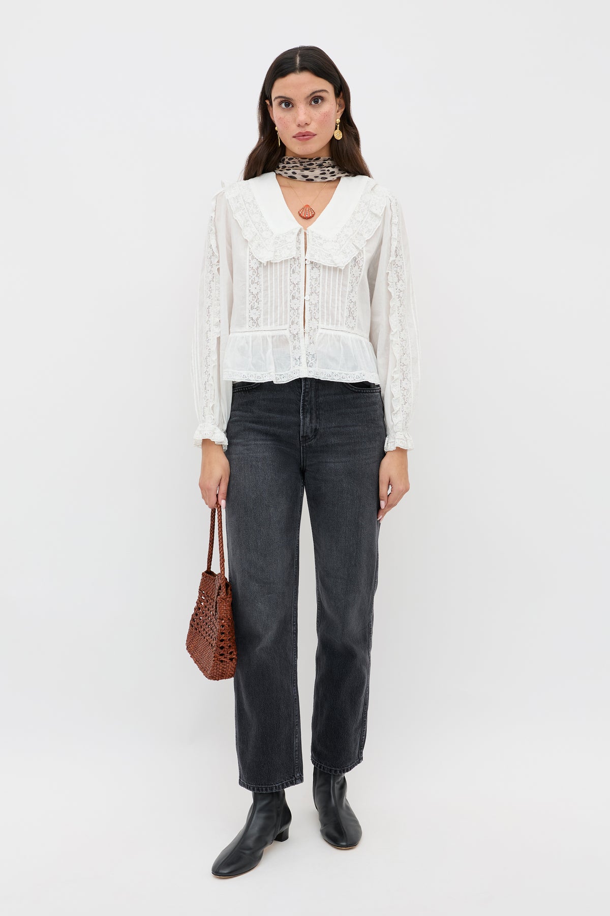 A woman stands against a white background wearing the Maribelle - Ivory vintage-inspired lace blouse with a large collar, styled with black jeans, black ankle boots, a patterned choker, pendant necklace, and a brown woven handbag.