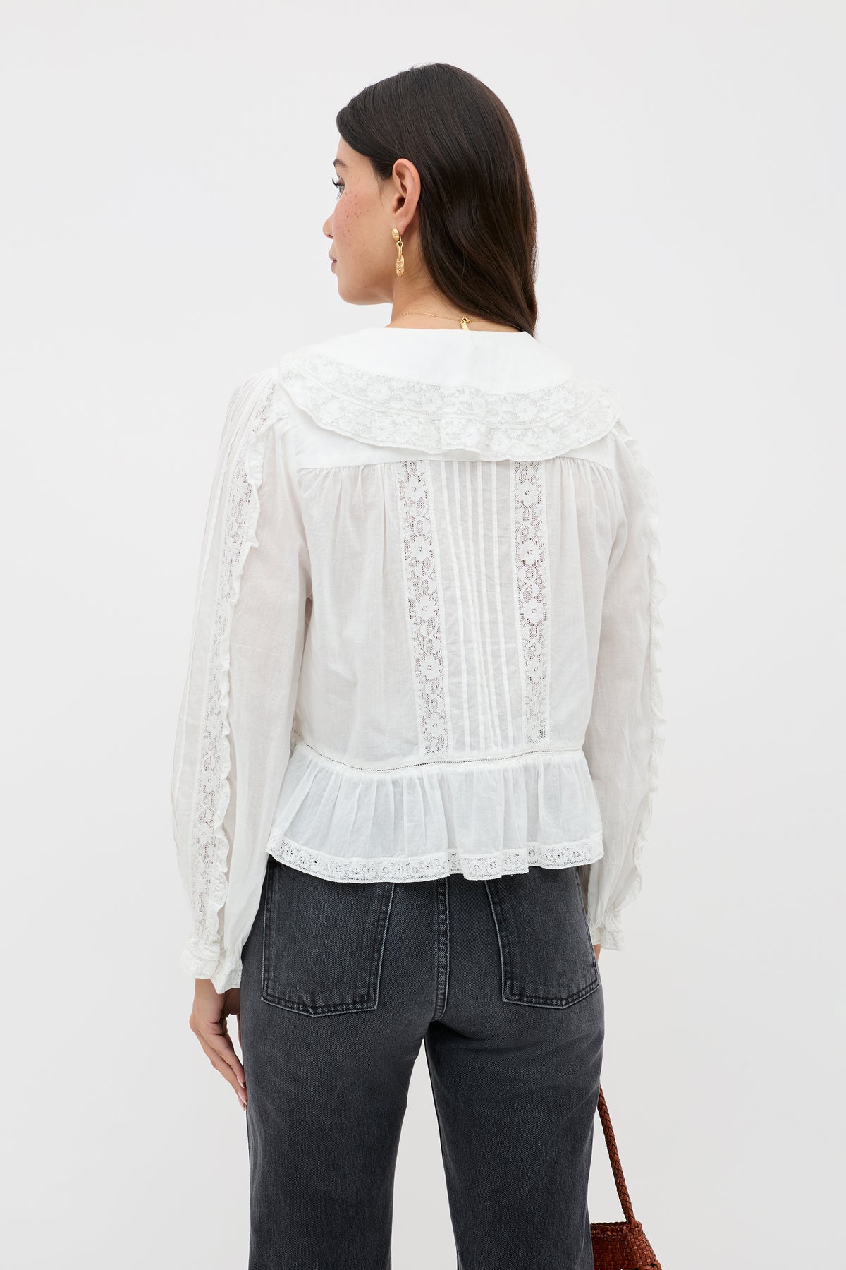 A woman with long dark hair, shown from the back, wears the Maribelle - Ivory blouse—a white vintage-inspired top with lace details, wide collar, and ruffled hem—paired with high-waisted black jeans and holding a brown bag.
