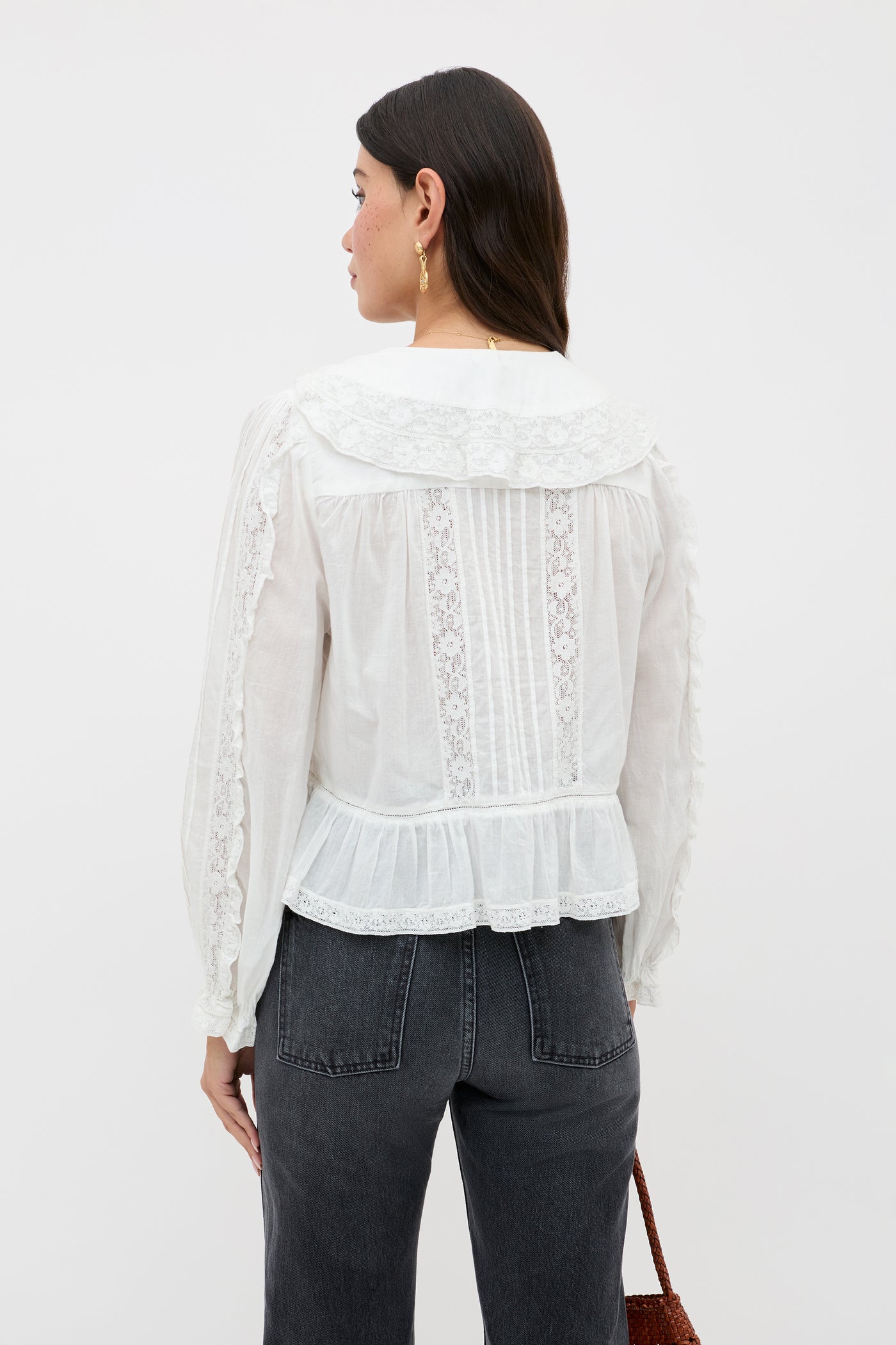 A woman with long dark hair, shown from the back, wears the Maribelle - Ivory blouse—a white vintage-inspired top with lace details, wide collar, and ruffled hem—paired with high-waisted black jeans and holding a brown bag.