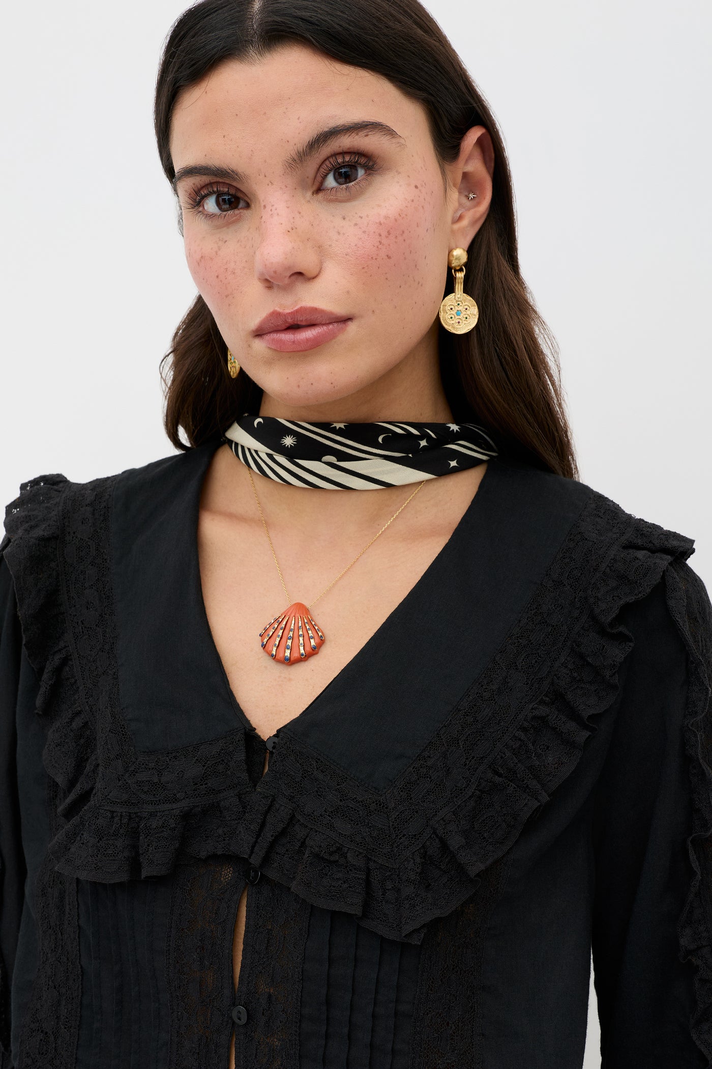 A woman wears the Maribelle - Black vintage-inspired ruffled blouse with a relaxed fit, paired with a black-and-white scarf, gold earrings, and a gold necklace with a red seashell pendant, set against a plain light background.