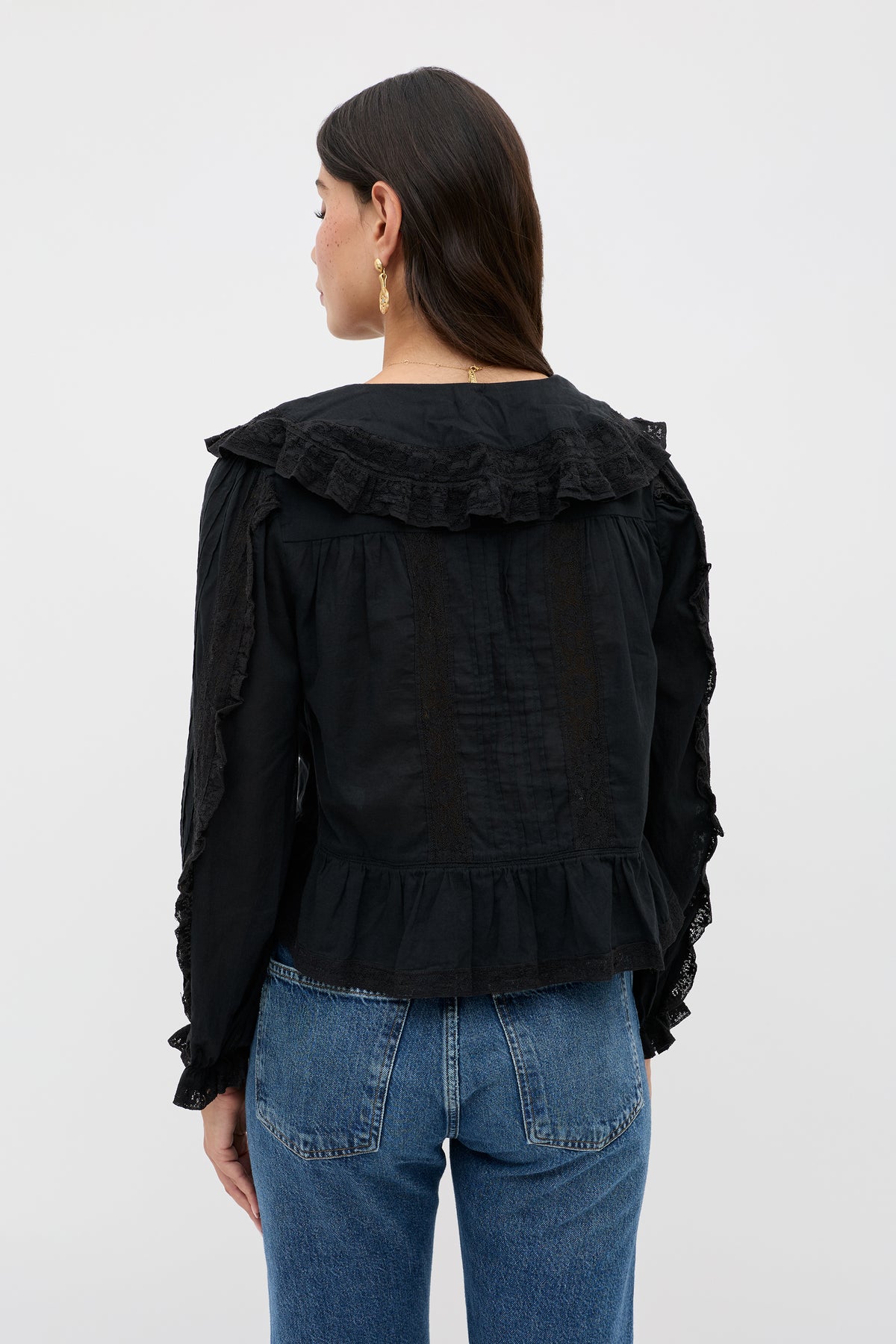 A woman with long dark hair is seen from the back, wearing Maribelle - Black—a vintage-inspired ruffled black blouse with lace accents and a peplum hem—paired with blue jeans and small gold hoop earrings.