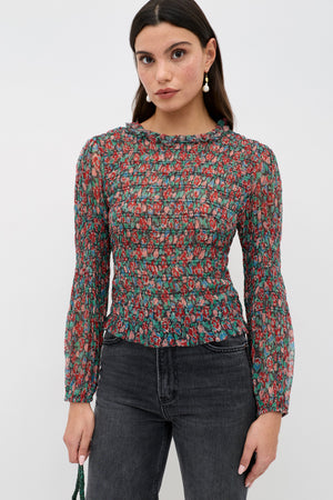 A woman wears the Gardenia - Jeannie Bloom Charcoal sheer, long-sleeve floral blouse with black jeans and drop earrings, standing against a white background and looking at the camera with a neutral expression.
