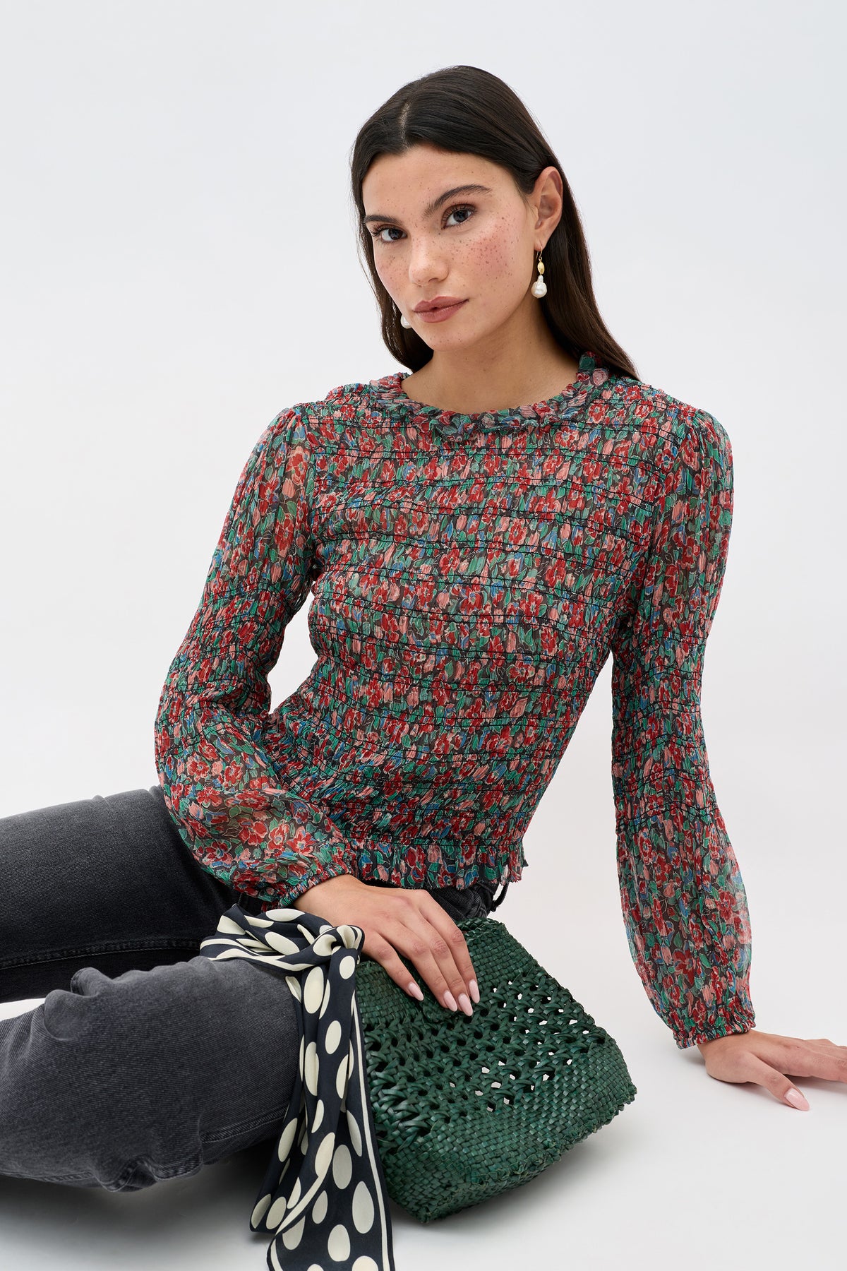 A woman with long dark hair sits on the floor in a sheer floral blouse, dark jeans, and drop earrings, holding the Gardenia - Jeannie Bloom Charcoal bag adorned with a black and white polka dot scarf against a light background.