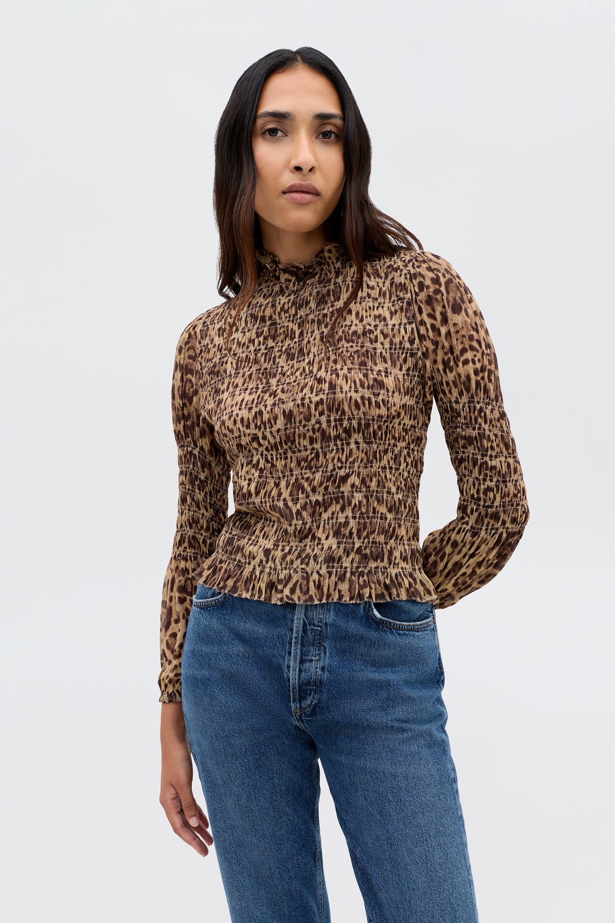 A woman with long dark hair wears the Gardenia - Mila Leopard Brown, a vintage-inspired fitted leopard-print top with ruffle trim, paired with blue jeans against a plain light gray background.
